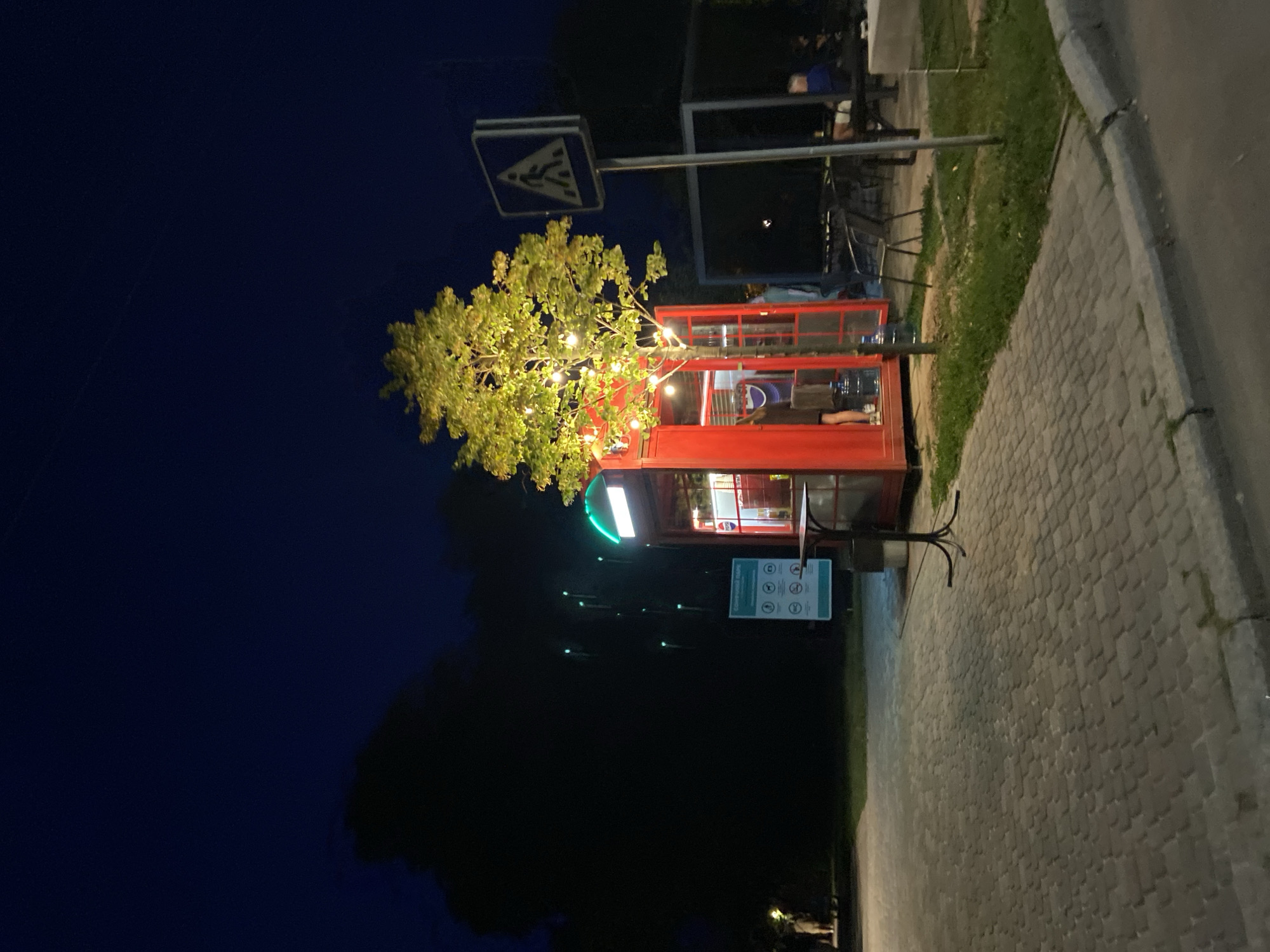 A red telephone booth, converted into a small kiosk, is situated on a cobblestone pavement at night. The kiosk is illuminated by string lights wrapped around a nearby tree. To the right of the kiosk, a pedestrian crossing sign is visible on a metal pole. In the background, there is a dark, shadowy area with some faint green lights, possibly a park or a wooded area. Further to the right, a covered seating area with tables and chairs is occupied by a few people. The overall scene suggests an outdoor area in Poltava, Ukraine, possibly a park or a public square, where the kiosk serves as a point of sale or service.