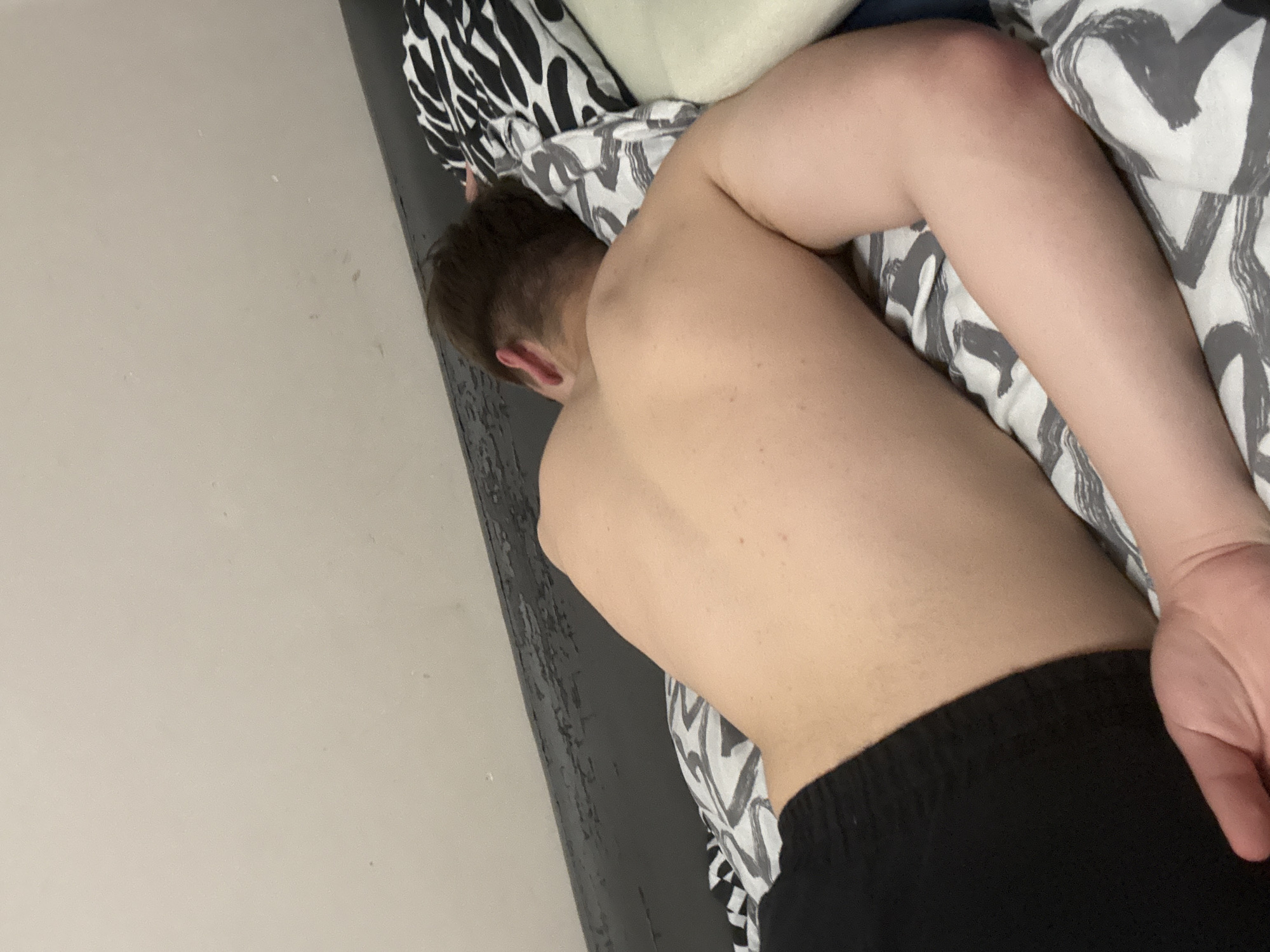 An indoor scene, likely a bedroom, is depicted from a slightly elevated viewpoint. A single individual, appearing to be a young adult male, is positioned on a bed. The person is shirtless, wearing black shorts or underwear, and lying on their left side with their back towards the viewer. Their head is turned away, resting on a pillow, indicating a state of rest or sleep. The individual's hair is dark and short.
The bed is covered with bedding featuring a distinct black and white geometric pattern. A pillow with this same pattern supports the person's head, and a second, lighter-colored pillow is partially visible to the right. Behind the bed, a dark-colored structure, possibly a headboard or part of a bed frame, exhibits a textured or worn surface. A plain, light beige wall with minor blemishes is visible above this structure, forming the background. The specific urban location of Sofia, Bulgaria, cannot be confirmed by the visual evidence within the image.