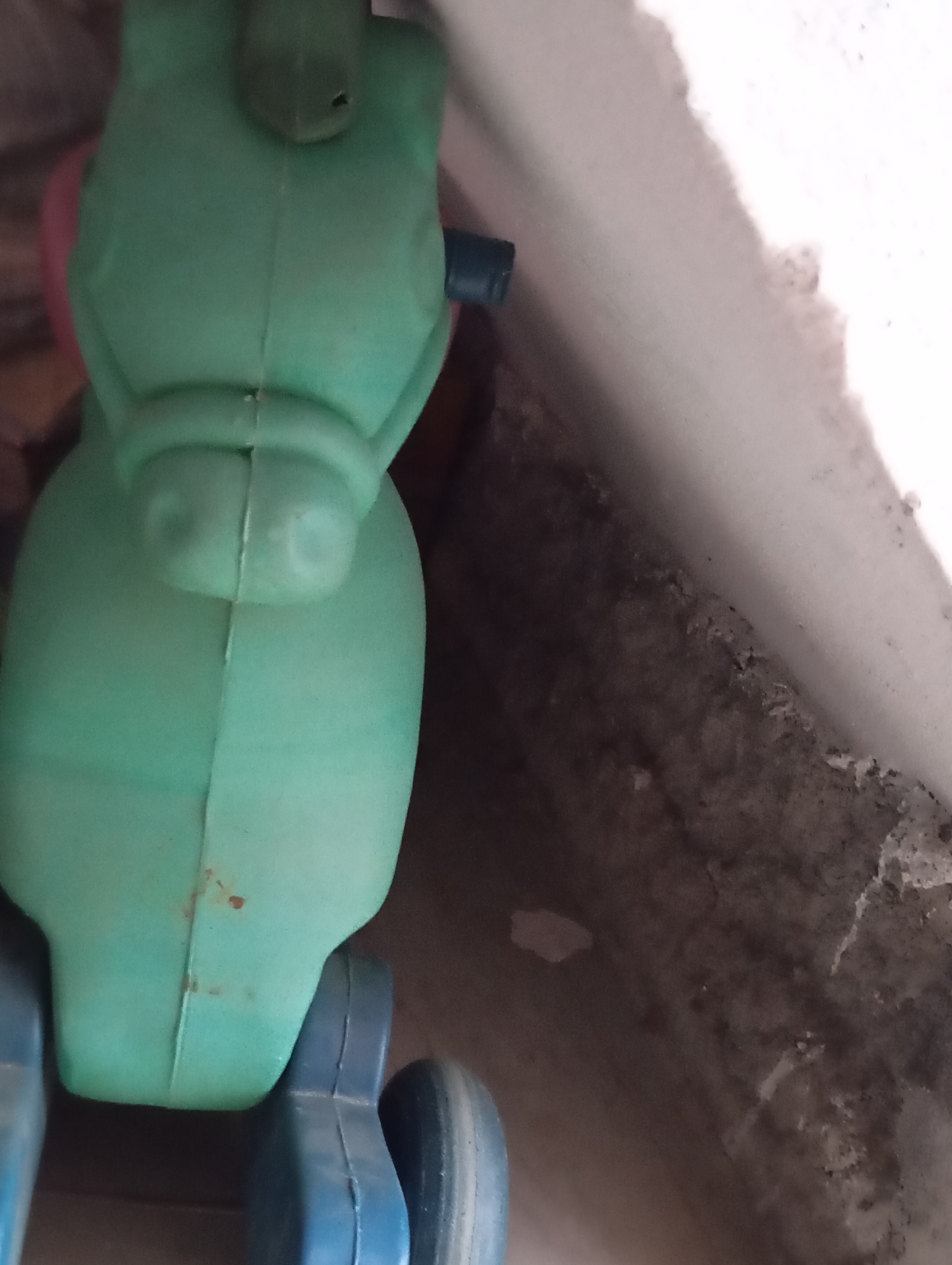 A close-up, overhead shot focuses on a green plastic toy, possibly a ride-on animal. The toy is largely obscured by the frame, with only the rear portion and wheels visible. The green plastic has a matte finish and shows some scuff marks and a reddish stain on the left side. Attached to the underside are two blue plastic wheel assemblies, each with a single wheel. The right wheel assembly is partially visible. The toy is positioned on a dark, rough surface with a white, textured edge to its right, suggesting an outdoor or utilitarian setting, perhaps a neglected corner of a yard or balcony. The lighting is diffused, casting soft shadows.