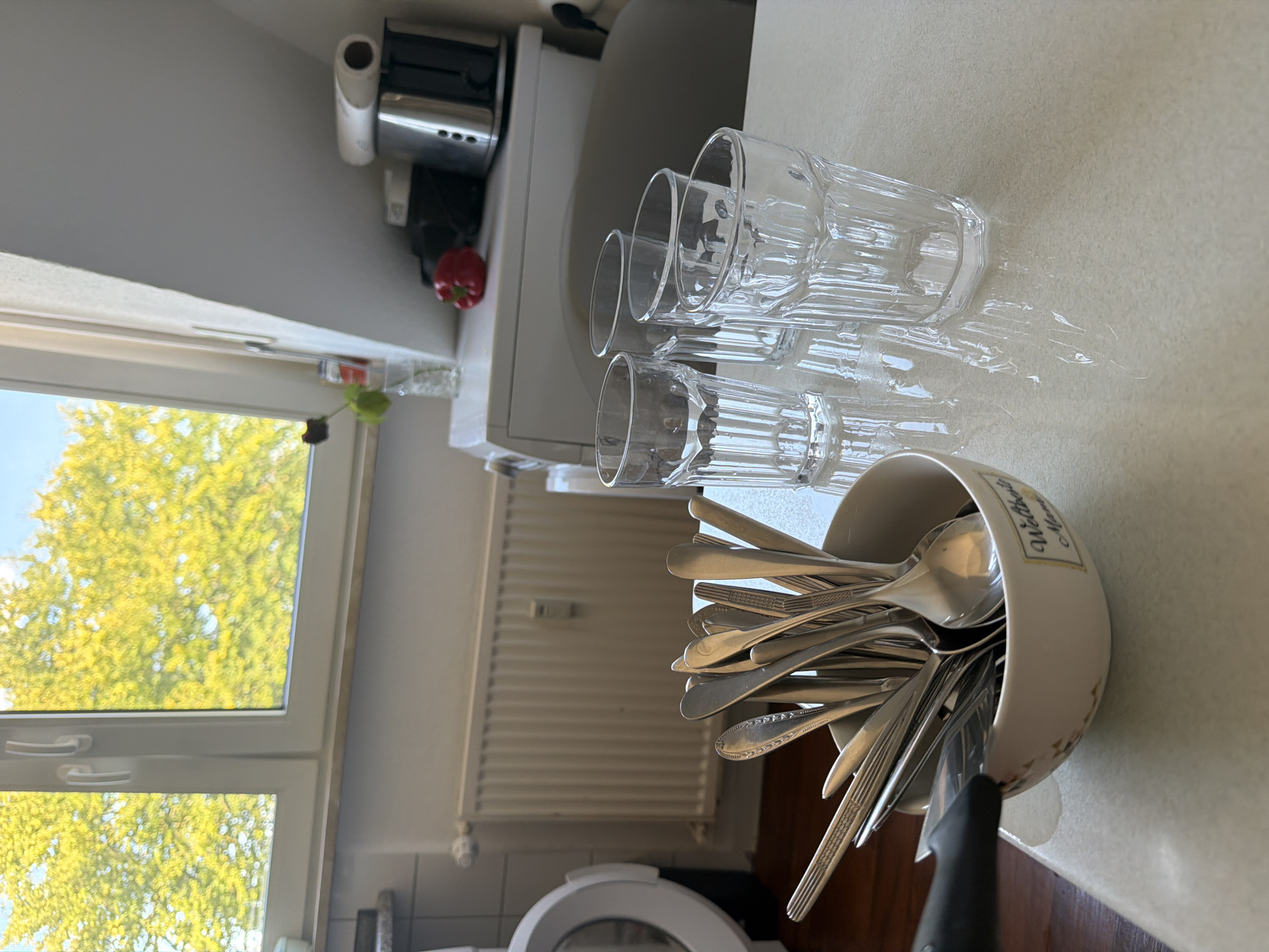 The image captures a domestic scene, likely a kitchen or dining area, with a window overlooking lush green trees. In the foreground, a bowl brimming with silverware, including spoons and forks, sits on a white tabletop. To the right of the bowl are four clear glasses, three of which are filled with water and reflect the light. In the background, a white washing machine is partially visible, with a red bell pepper and a toaster on a shelf above it. The overall impression is one of a well-lit, everyday environment. The specific location in Nienburg/Weser, Germany, cannot be determined from the visual information provided.