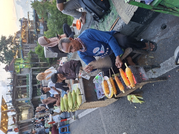 The scene is a street food vendor in Kecamatan Piyungan, Indonesia (latitude: -7.846, longitude: 110.48). A man is grilling corn on a charcoal grill, while another man sits on a wooden stool to his right. The man is using tongs to turn the corn, which is on skewers. There are several other people in the background, including two women wearing headscarves, and a young man wearing a white shirt and black pants. There are bags of corn stacked on a wooden table beside the grill. The scene is set on a paved road with a concrete curb. The background shows several buildings and trees.