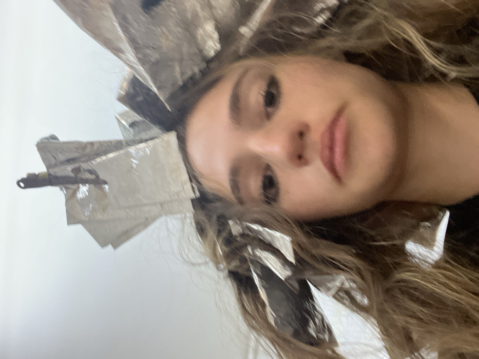 The image features a young female subject, facing forward, with her head tilted slightly upwards towards the camera. Her light brown hair is extensively sectioned and wrapped in numerous pieces of reflective aluminum foil, indicating a hair highlighting or coloring process is underway. A single dark metal clip is visible near the top center of her head, securing some of the foils. Her eyes are open, and she has a neutral facial expression with naturally colored lips. Her skin tone is fair. Only the upper part of her torso is visible, clad in a dark, indistinct garment. The background is uniformly light-colored and out of focus, suggesting an indoor setting such as a hair salon. No identifying features of a specific city or location, including Puebla, Mexico, are discernible within the image.