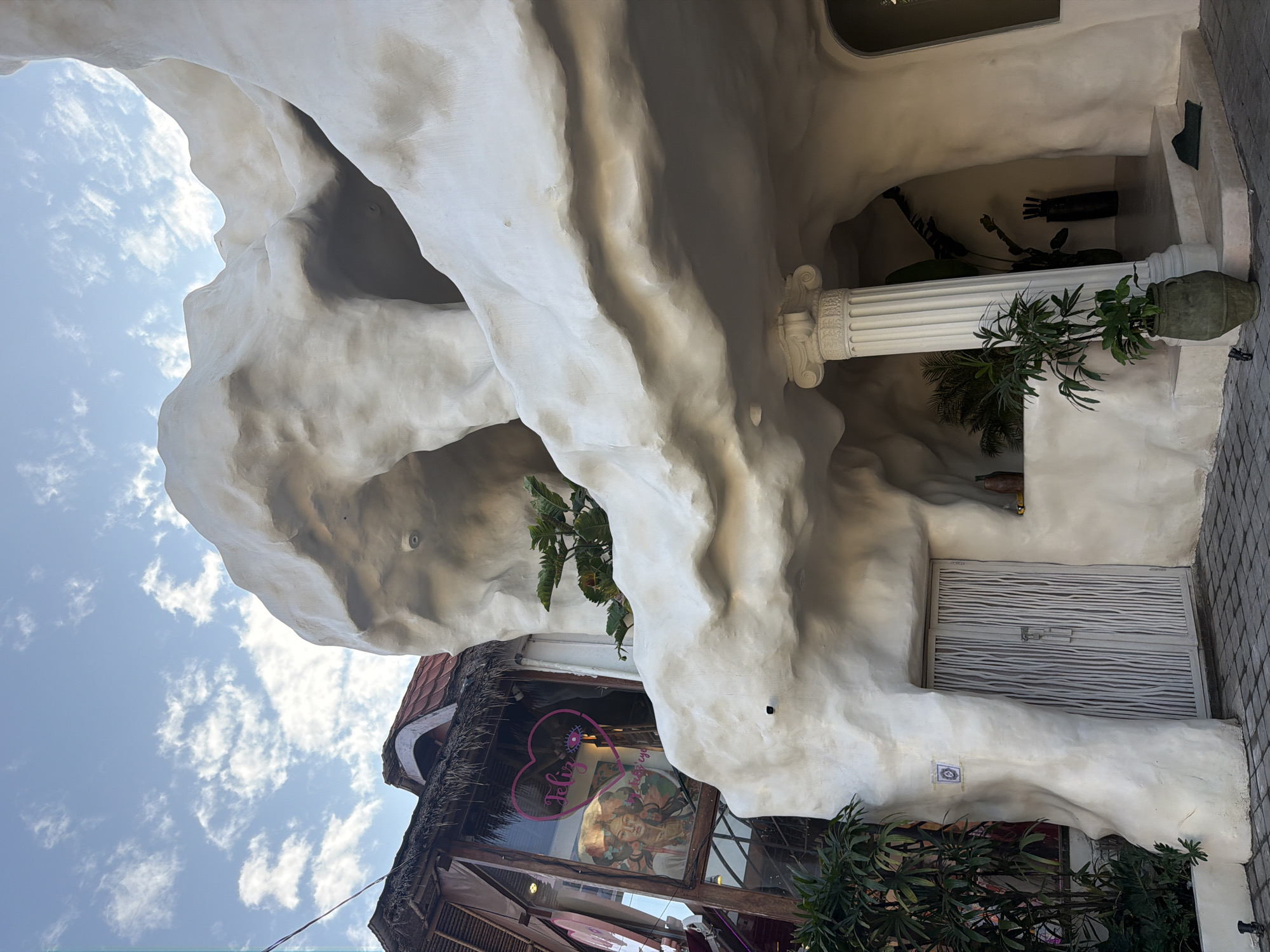 The image depicts the exterior of a building with a unique, organic, cave-like architectural design. The structure is predominantly white and appears to be made of a textured material, possibly stucco or concrete, molded into rounded, flowing shapes that resemble natural rock formations. In the foreground, a cobblestone pathway leads to a white door with vertical slatted panels, flanked by curved, wall-like structures. To the left of the door, a lush green plant in a dark pot is situated, with more greenery visible behind it and to the left, against a building with a thatched roof and glass windows. Through one of the windows, a vibrant artwork of a woman with elaborate headwear is visible, along with a neon pink heart-shaped graphic and text that reads "Feliz eye".
To the right of the entrance, a grand Corinthian-style column supports an overhang of the sculpted white architecture. Behind the column, steps lead up to an arched opening, revealing an interior space with more plants, including large-leafed tropical varieties, and decorative dark vases. The scene is set against a bright blue sky with scattered white clouds, suggesting daytime. The overall impression is one of an artfully designed establishment, possibly a boutique, cafe, or gallery, blending naturalistic elements with a modern aesthetic. The specific location is identified as the Ubud District, Indonesia, a region known for its artistic and cultural heritage.