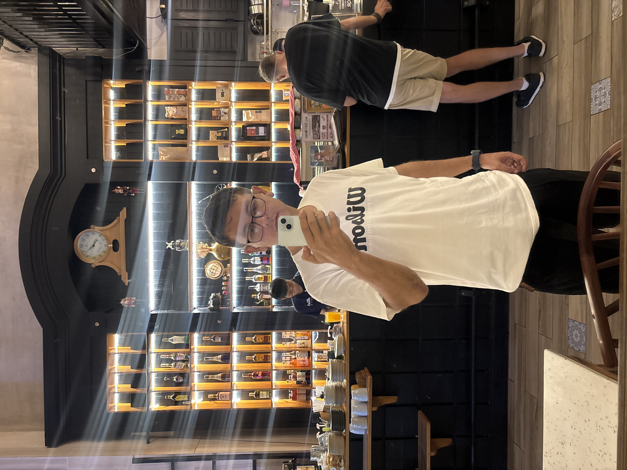 This image captures a man taking a photograph of himself in a reflective surface, likely a mirror, within an indoor establishment in Unknown, Unknown. The man, positioned centrally, wears glasses, a white t-shirt with the reversed word "Wilsou" (suggesting "Wilson"), black trousers, and a dark smartwatch on his left wrist. He holds a light green smartphone to his face.
Behind him, in the reflection, is a bar or service counter area. A male staff member in a dark blue polo shirt is visible on the left, working behind the counter with a glass containing an orange beverage nearby. To the right, another male individual, wearing a black t-shirt, beige shorts, and dark sneakers with white socks, stands with his back to the viewer, leaning on the counter.
The background wall features elaborate dark-colored shelving units with integrated warm LED strip lighting. The upper section includes a decorative arch containing a wooden mantel clock and a nutcracker figurine. The shelves display various items: liquor bottles are prominent on the left side, while the right side holds packaged goods (possibly coffee), a decorative globe, a vintage camera, small figurines, and a gold-colored vase. The dark counter below is laden with stacks of white and dark plates, cups, and other service items.
The floor consists of light-colored wooden planks, with two square blue and white patterned tiles embedded within. A corner of a light-speckled table and part of a wooden chair are visible in the bottom foreground. Strong horizontal and diagonal light streaks are present across the image, indicative of reflections from a light source off the reflective surface.