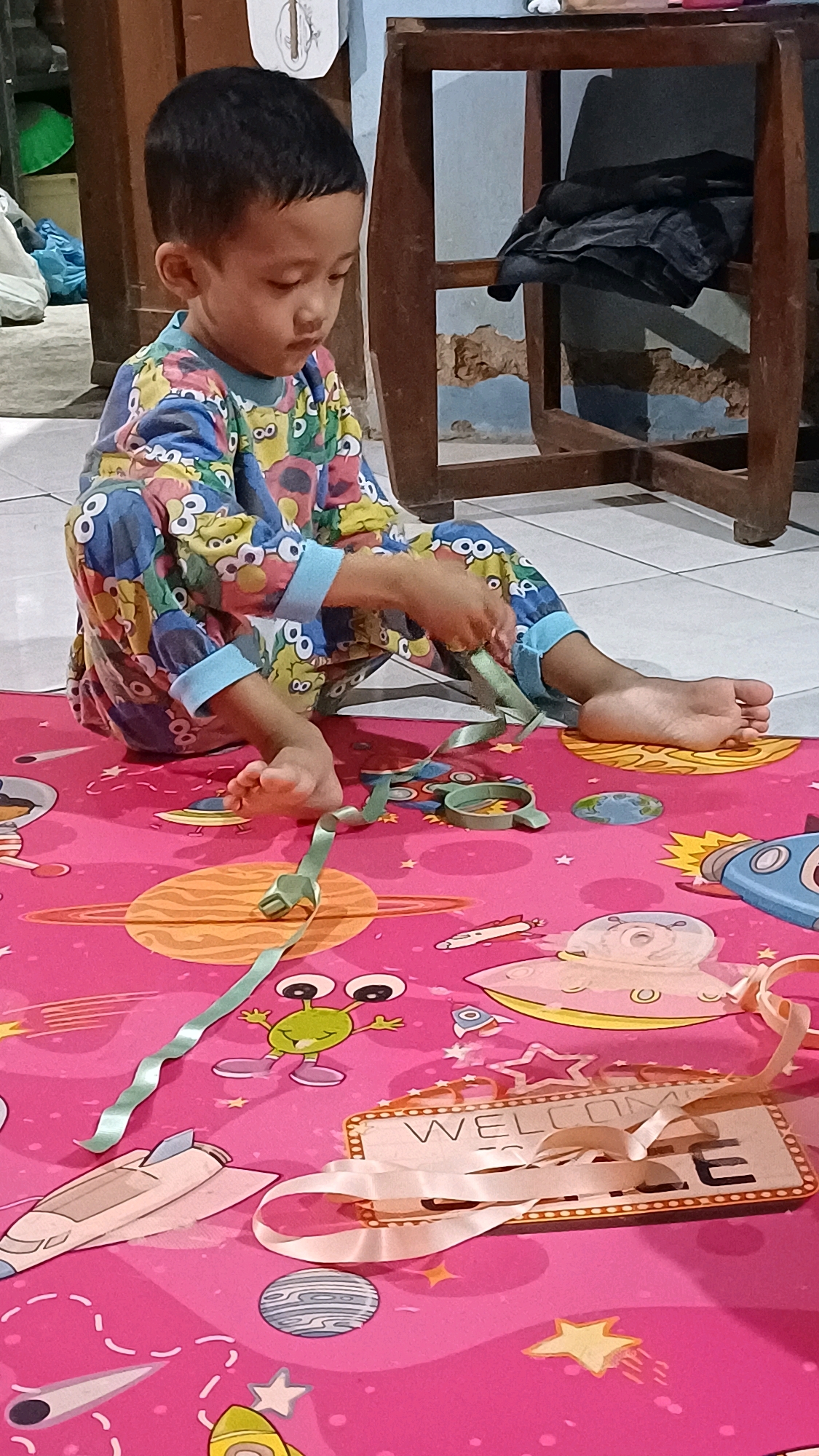 A young child, likely of Indonesian descent, is seated on a vibrant pink playmat adorned with space-themed graphics, including planets, rockets, and aliens. The child is wearing pajamas with a colorful, cartoonish pattern featuring various characters, possibly from a children's show. The child's hands are engaged with a thin, light green ribbon, manipulating it as if playing or creating something. The playmat is spread on a tiled floor, characteristic of an indoor setting. In the background, a wooden piece of furniture with distressed paint is visible, along with some fabric items. The lighting suggests an indoor environment, possibly a home. The scene appears to be set in Wonokerso, Indonesia, based on the provided location context.