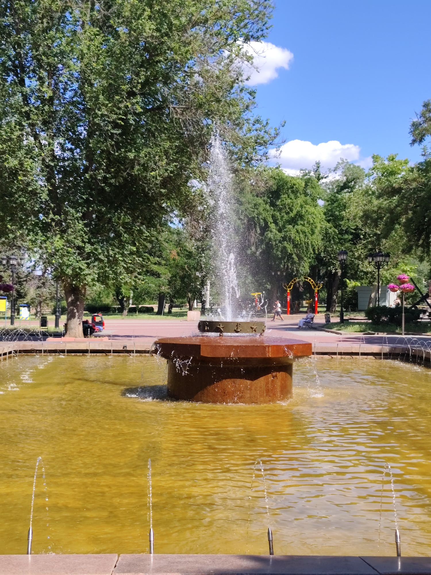 This is a photograph of a fountain in a park. The fountain is in the foreground, with water spraying upwards from a central structure. The water in the fountain pool is a murky yellow color. In the background, there are trees and some people. The sky is blue with some white clouds. The fountain is located in the City Garden in Mykolaiv, Ukraine.