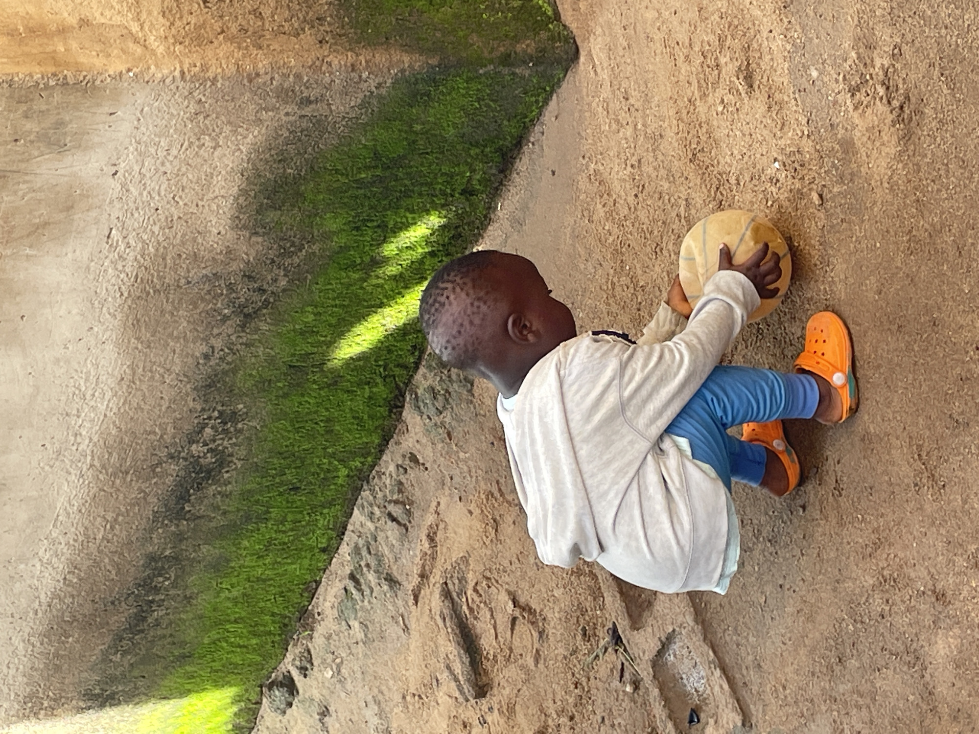 In Barakin Lerawi, Nigeria, a young child, possibly a toddler, is crouched down on sandy ground. The child is wearing bright orange Croc-style shoes, light blue pants, and a light-colored hooded jacket. The child is interacting with a ball, which appears to be made of a light-colored, textured material, possibly a worn basketball. The background consists of a rough, textured wall with patches of vibrant green moss, particularly on the left side, illuminated by bright sunlight. The ground is composed of loose sand and dirt, with some embedded stones and debris. The overall scene suggests an outdoor environment, likely a yard or alleyway, common in residential areas of Nigerian cities.