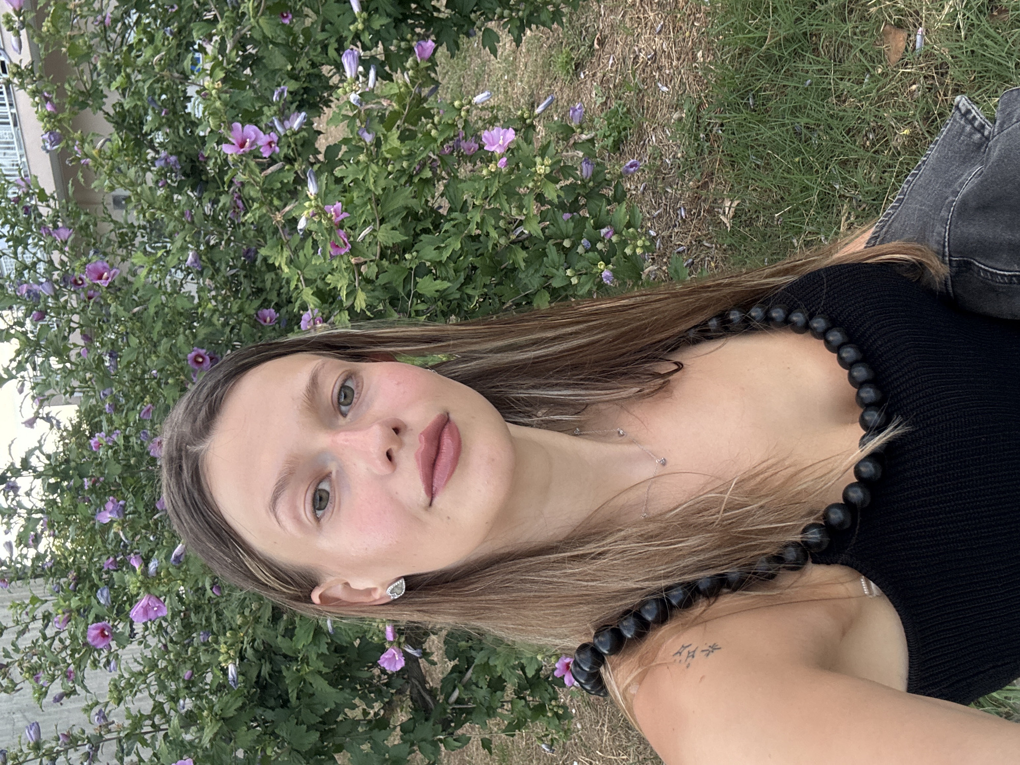 A young woman with shoulder-length brown hair is photographed outdoors, in Stavros, Greece. She is wearing a black top with a beaded neckline and a small silver necklace. She has a tattoo on her left shoulder. The background consists of a large green bush with purple flowers. The ground is covered with dry leaves and grass. The image is a close-up, with the woman facing the camera and smiling.