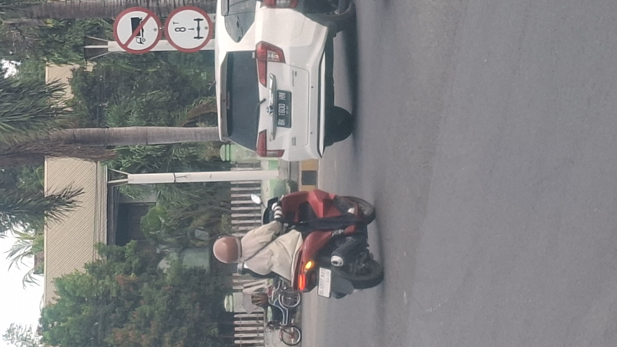 The image captures a street scene, possibly in Lhokseumawe, Indonesia, featuring a person riding a red scooter and a white car. The scooter rider, facing away from the camera, wears a light-colored long-sleeved shirt, dark pants, and a light brown helmet. A dark bag is slung across their chest. The scooter appears to be a modern automatic model with its taillights illuminated. To the right of the scooter, a white car is visible, with its rear bumper and license plate, which reads "BK 1800 HM," clearly displayed. The car is positioned in what appears to be a traffic lane.
In the background, a white fence with vertical wooden posts lines the edge of the road. Beyond the fence, there are lush green trees and the roof of a building. Traffic signs are visible on a pole; one is a circular red sign with a diagonal line and an image of a truck, indicating "no trucks," and below it, another circular sign with a red border shows "8 T" with an arrow pointing down, likely indicating a weight limit for vehicles. Palm trees are also present, suggesting a tropical or subtropical climate. The road surface is asphalt, and the lighting suggests it is daytime, possibly late afternoon or early morning due to the soft, diffused light. The overall scene is one of everyday urban traffic and infrastructure.