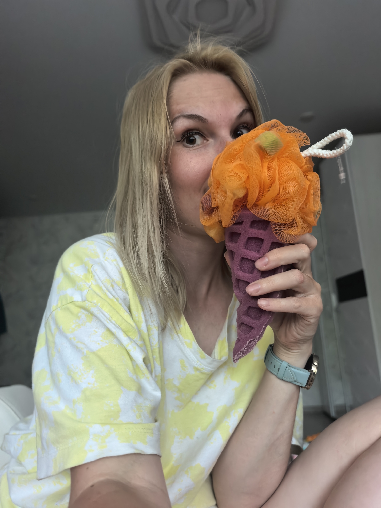 A woman with blonde hair and a yellow and white tie-dye shirt is holding a bath sponge designed to look like an ice cream cone in front of her face. The "ice cream" part of the sponge is orange and poofy, while the "cone" is purple with a waffle pattern. She is looking at the camera with wide eyes. She is wearing a watch with a light blue band. The background appears to be the interior of a home, with a neutral-colored wall behind her. The image does not include any details that would allow identification of the location as Sterlitamak, Russia.