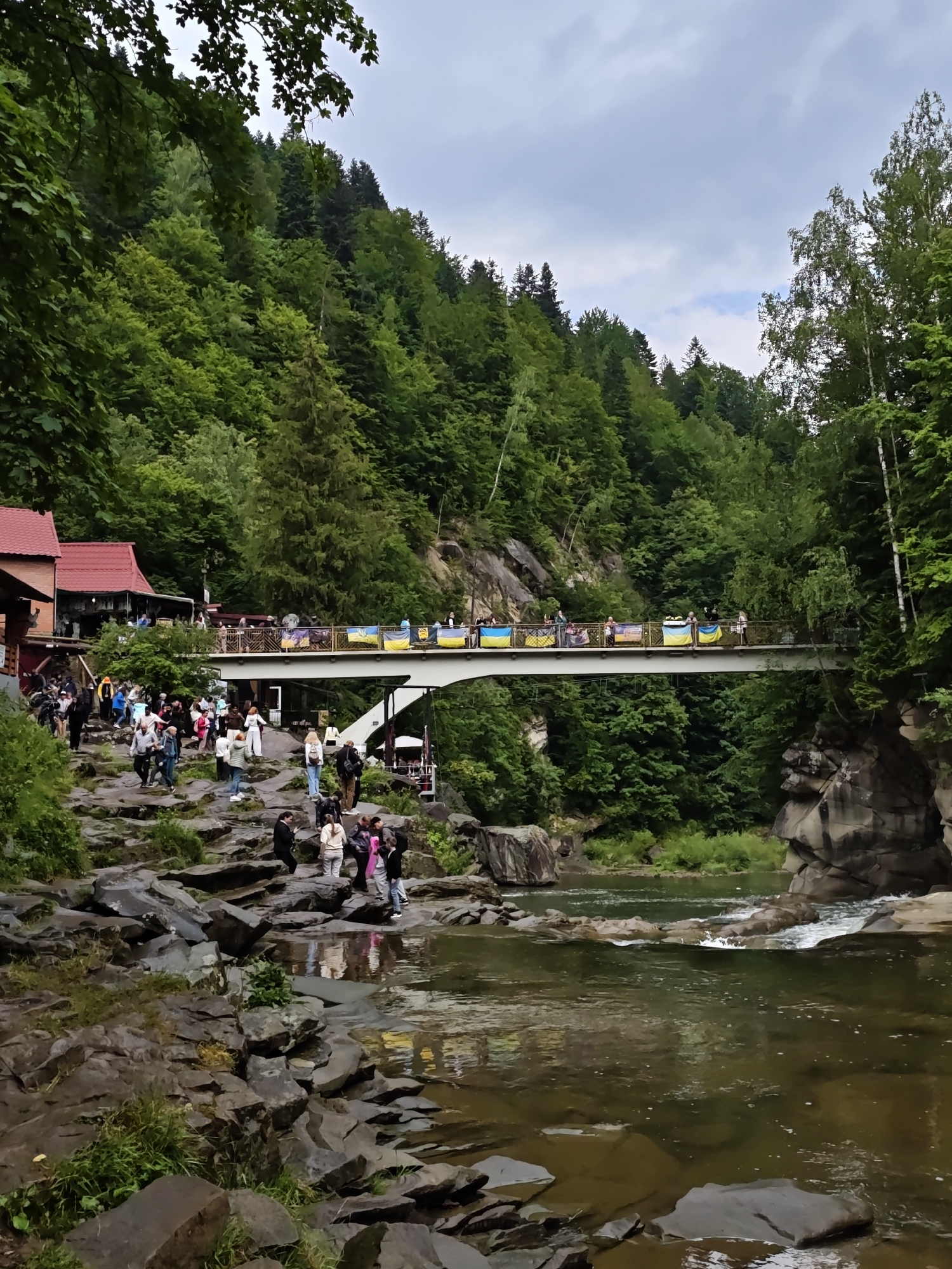 This image captures a view of the Prut River in Yaremche, Ukraine, with a pedestrian bridge spanning across it. The bridge is decorated with Ukrainian flags and banners, with numerous people walking across it and on the rocky riverbanks. Lush green trees cover the steep hillsides surrounding the river. A building with a red roof is visible on the left bank, and the river flows over rocks, creating a scenic, natural environment.