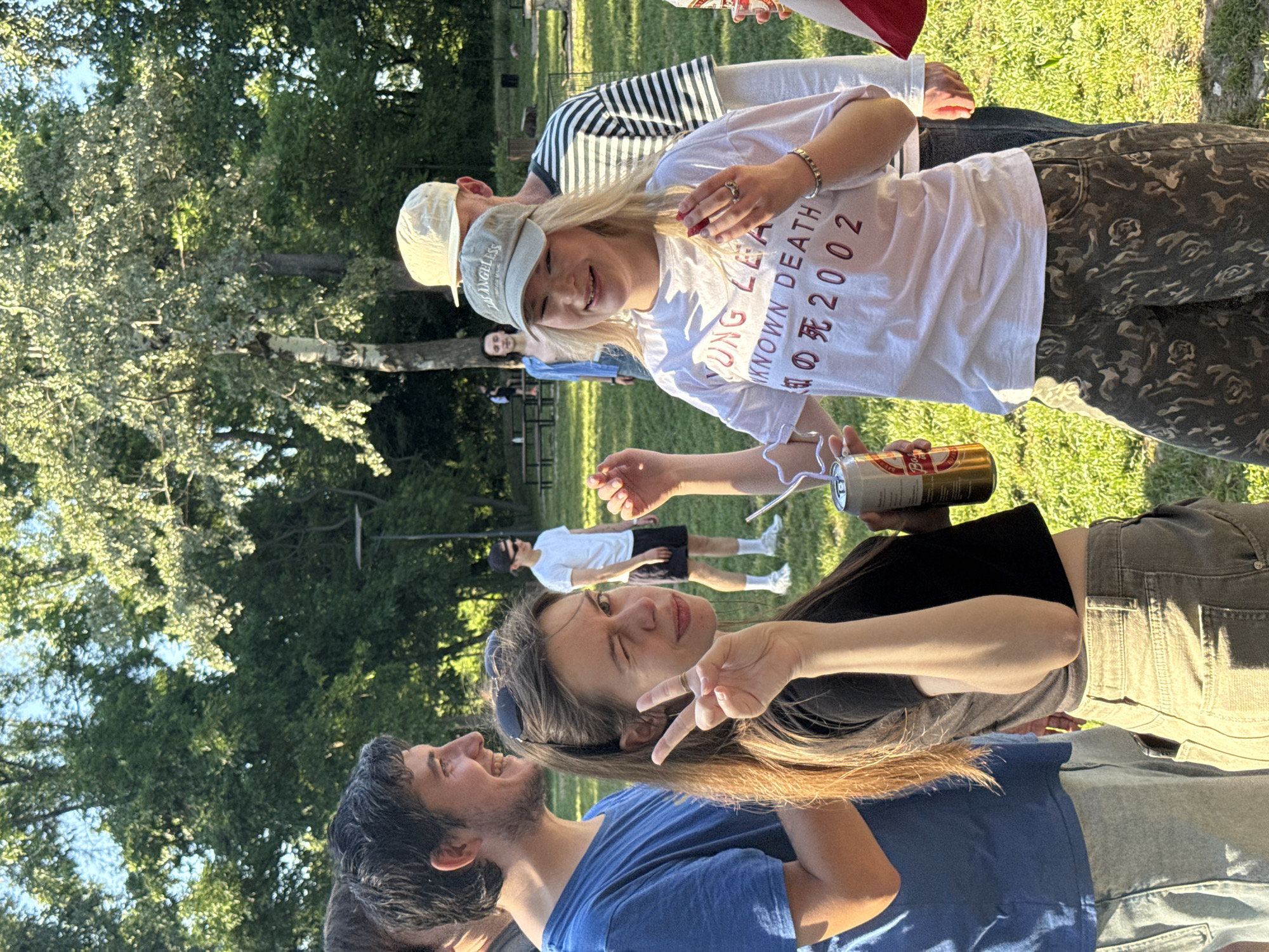 A group of people are gathered outside in a grassy area, likely a park. In the foreground, a young woman wearing a black top makes a peace sign towards the camera while a man in a blue shirt laughs beside her. A blonde woman in a white shirt is holding a can of whipped cream, and appears to be squirting it. A can of beer is in her other hand. Other individuals are visible in the background, including one wearing a striped shirt, as well as other people indistinctly. The environment appears to be outdoors, with trees and grass, suggestive of a park setting. It is impossible to determine whether the picture was taken in Cukarica Urban Municipality, Serbia.