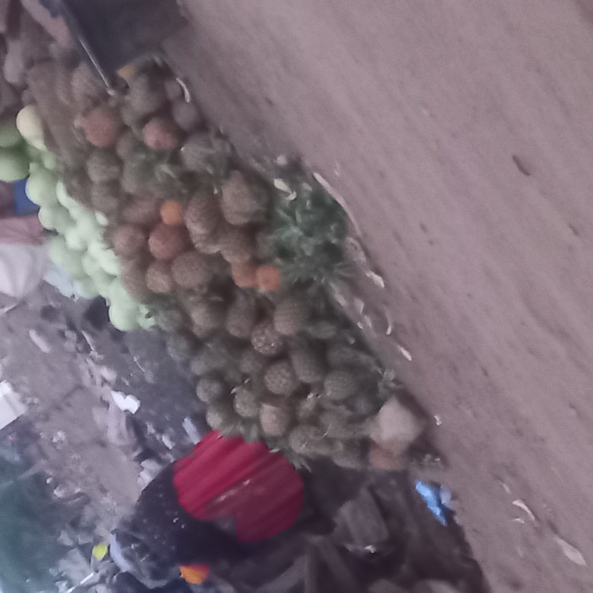 The image appears to be taken from an elevated perspective in Barnawa, Nigeria. A vendor with a red head covering is shown near a display of produce. The produce includes pineapples, some appearing green and unripe, others with a more mature brown coloration, and a separate collection of green, spherical fruits, potentially limes. The produce is arranged near what appears to be a concrete or stone surface. Other items and debris are visible in the lower left quadrant.