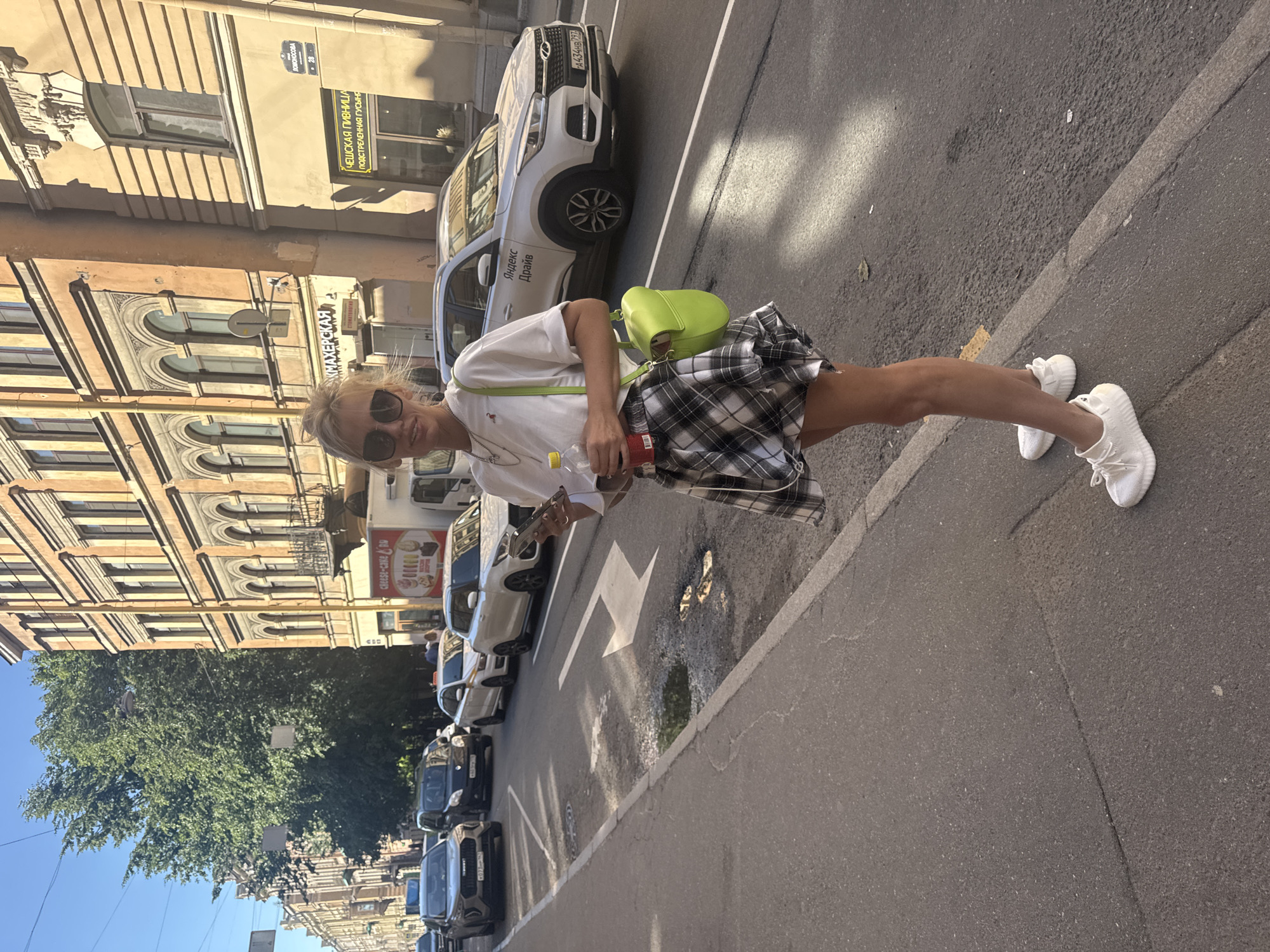 A blonde woman with sunglasses, holding a phone and a bottle of red liquid, stands on a city sidewalk. She is wearing a white t-shirt, a black and white checkered skirt, and white sneakers. A bright lime green crossbody bag is slung over her shoulder. Behind her, a street is lined with cars and multi-story buildings with arched windows. A white car with "Яндекс Драйв" visible on its side is parked in the street. The architecture suggests the location is Saint Petersburg, Russia. The street appears to be under a bright, sunny sky.