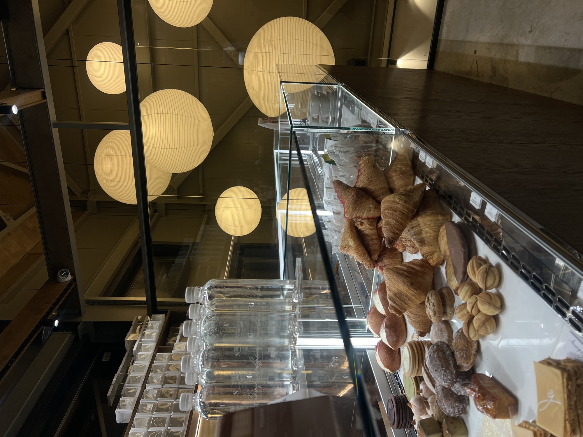 The image captures an interior scene in Vladivostok, Russia. Overhead, several spherical paper lanterns provide illumination. Below, a glass display case exhibits an array of pastries, including croissants, cookies, and cakes. Bottled water is visible in a shelf adjacent to the display. The environment suggests a bakery or cafe setting.