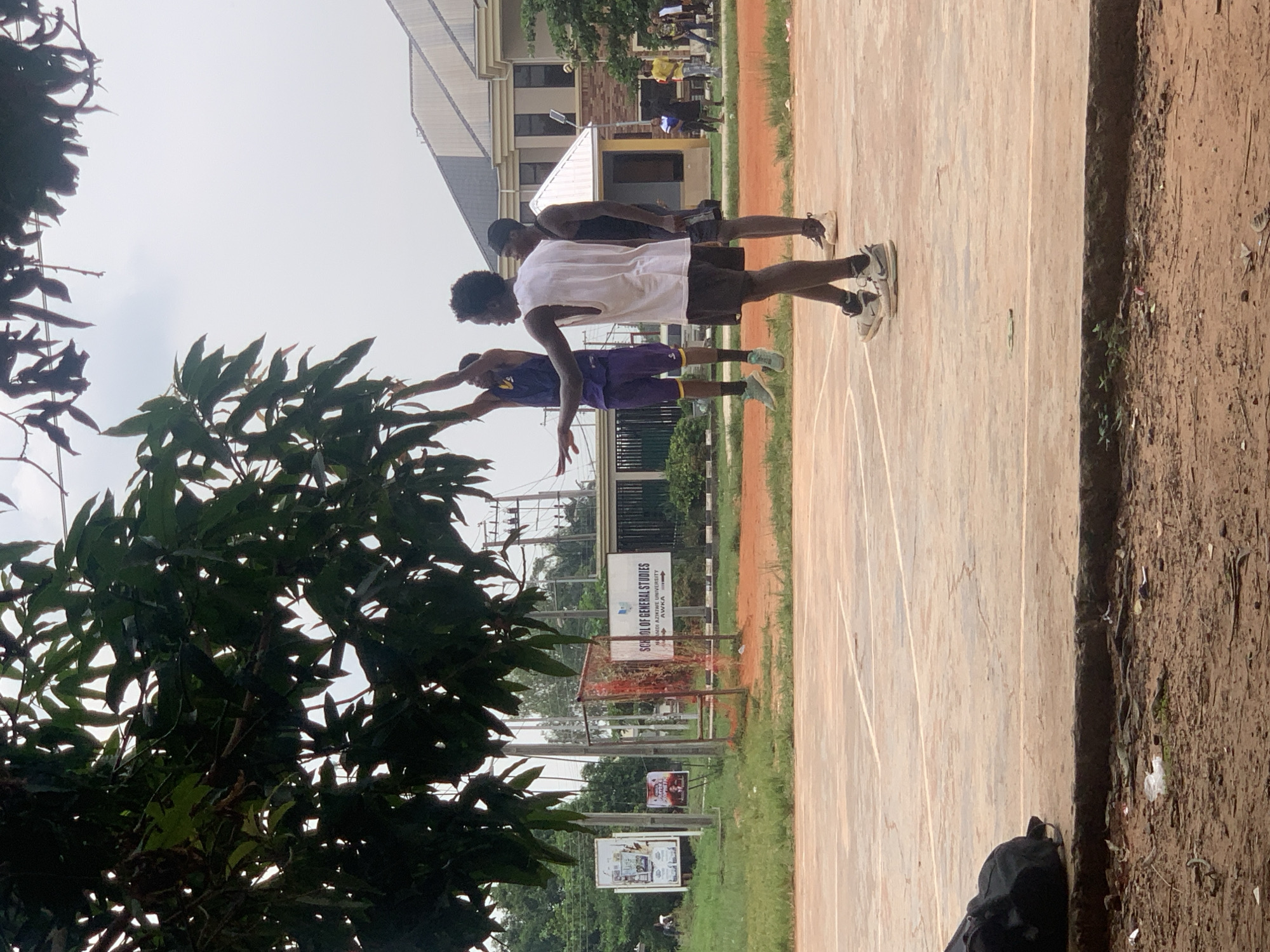 In Ukukwa, Nigeria, on an outdoor basketball court, three young men are engaged in a game. One player in a purple jersey and shorts jumps to shoot the ball, while another player in a white sleeveless shirt and black shorts stands nearby, seemingly observing the play or preparing to defend. A third player, wearing a black tank top and shorts, stands behind the player in white. The court's surface is a reddish-brown concrete, marked with white lines. Trees with dark green leaves overhang a portion of the court. In the background, a building with a light-colored facade and a sloped roof stands, and further beyond, there are more trees and electrical poles. Near the edge of the court, a black bag rests on the ground.