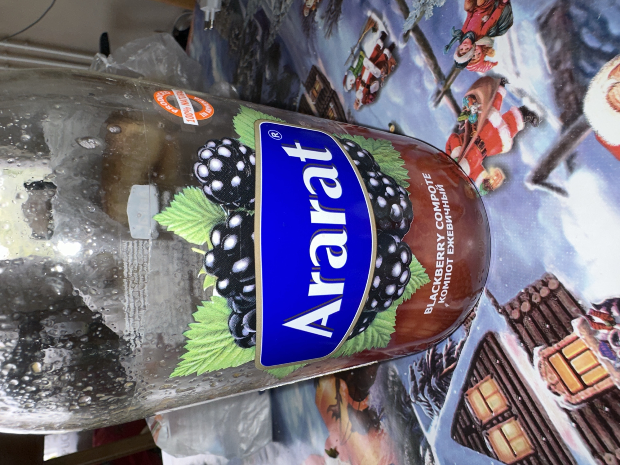 A close-up, slightly distorted, exterior shot shows a clear glass jar, partially filled with a dark liquid, sitting on a tablecloth adorned with a winter scene. The jar's exterior is covered with water droplets, suggesting it may have been refrigerated or recently washed. A label on the jar features a blue rectangle with the white, stylized text "Ararat" and a registered trademark symbol. Above this, an illustration of blackberries and green leaves is present. To the right of the label, a circular sticker reads "PRODUCED 100% NATURAL IN ARMENIA". The lower portion of the jar is a reddish-brown color, and the text "BLACKBERRY COMPOTE" and "КОМПОТ ЕЖЕВИЧНЫЙ" is visible. The tablecloth displays a snowy landscape with log cabins, illuminated windows, and figures of Santa Claus and children in winter attire. The background is dark and out of focus.