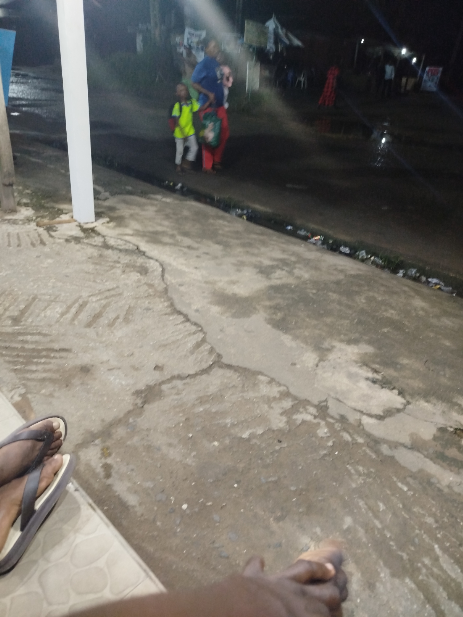 A low-angle, night-time photograph depicts a street scene in Ikot Mbonde, Nigeria. The foreground features cracked concrete, and the lower left corner shows a pair of feet clad in flip-flops. An arm rests on the concrete as well. In the middle ground, a street is visible, with trash accumulating along its edge. A woman carries a child in her arms, while another child walks alongside her, wearing a light-colored top and shorts. In the background, a group of people stand under a streetlight, and some red cloth is visible. A vertical white beam partially obscures the view to the left of the woman and child.