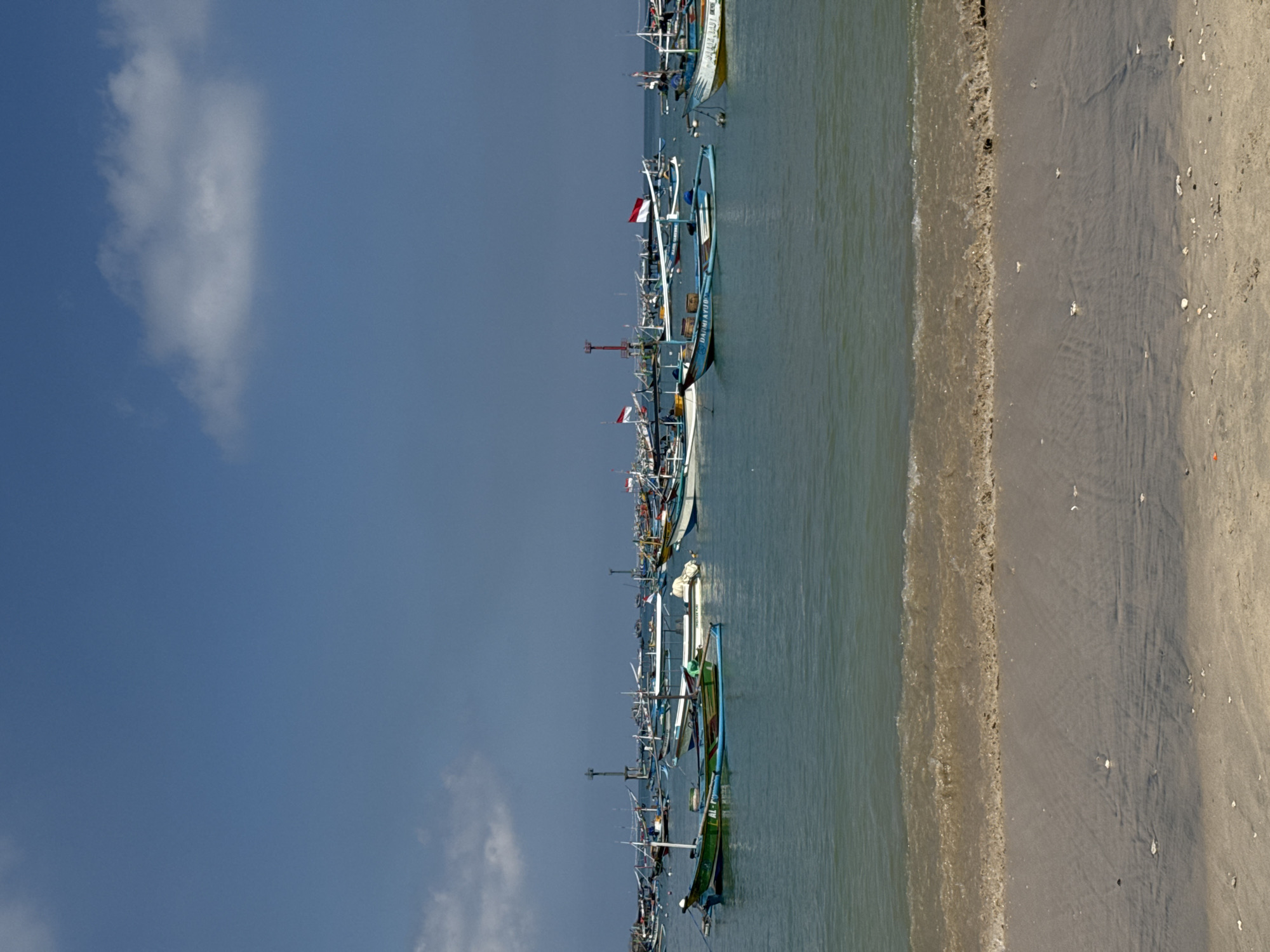 This image captures a tranquil daytime scene on a beach in Kuta, Indonesia, featuring a row of traditional fishing boats, calm waters, and a clear sky.
Along the shoreline, numerous colorful traditional outrigger fishing boats, known as jukung, are moored in the shallow water. These boats are varied in design and color, with prominent hues of light blue, white, green, and some with patterned hulls. Many of the boats prominently display red and white Indonesian flags on their masts, indicating their national origin. Text is visible on some vessels; for example, "DARMA AYU" is painted on the side of a light blue boat in the mid-ground, and "PUTRA LAUT" can be faintly discerned on a white and yellow boat further down the line.
The sea water is calm, appearing a muted greenish-grey, with gentle ripples reflecting the clear sky. It meets a sandy beach on the right side of the frame. The beach sand is a light brown color, marked by subtle texture and scattered small shells or debris, with a narrow band of frothy white waves at the water's edge.
The sky, occupying the upper left portion of the image, is a vibrant blue, largely clear with only a few soft, wispy white clouds. The bright, even lighting suggests it is a clear day, likely morning or afternoon. The overall scene depicts a typical coastal setting with local fishing activity in a serene environment.