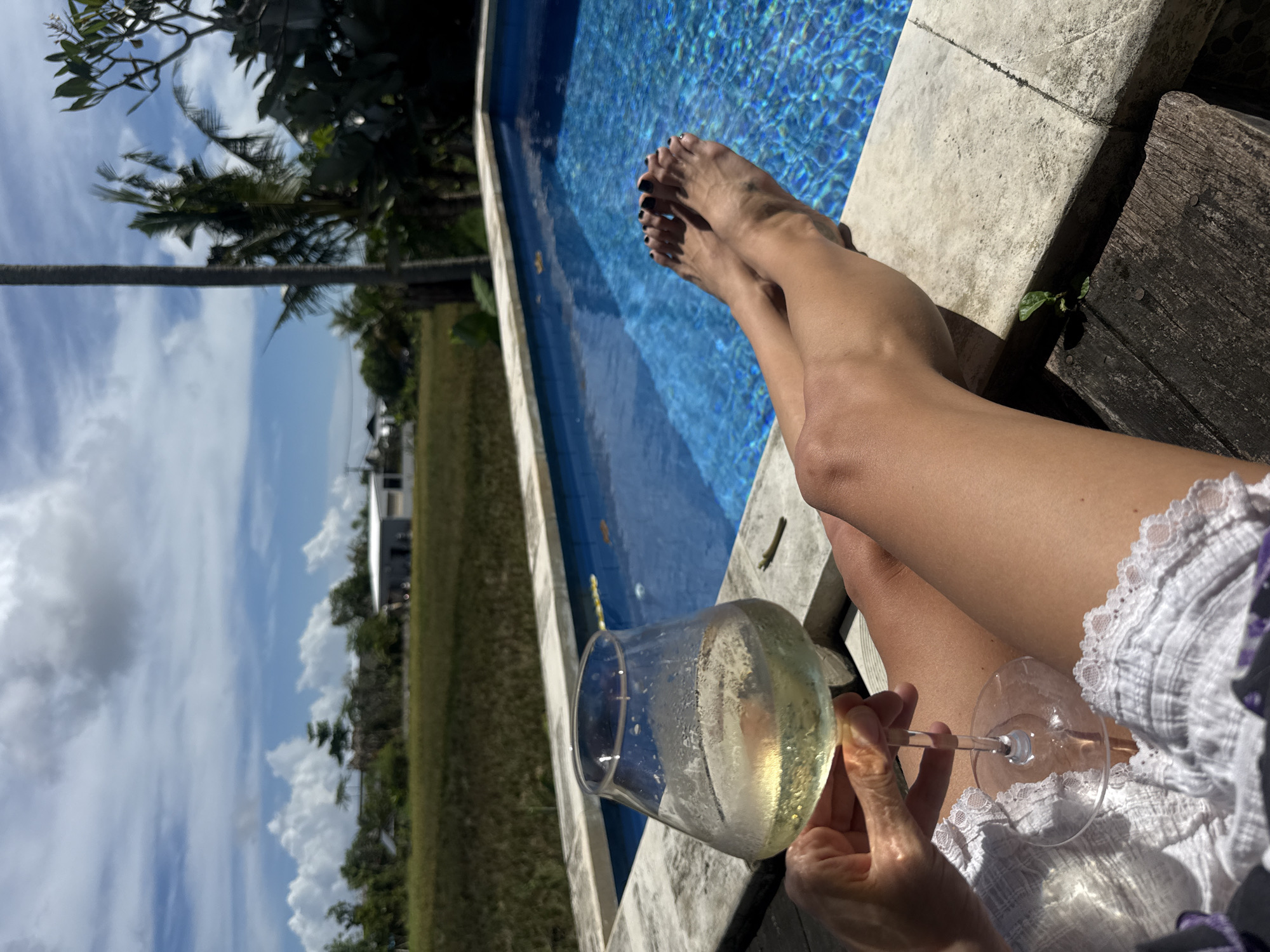 The image shows a person relaxing by a swimming pool. The person's legs are extended towards the pool, with their feet visible near the edge. They are holding a glass of white wine. The pool has clear blue water and is bordered by a light-colored stone edge. In the background, there is a grassy area with some buildings and trees under a partly cloudy sky. The location is Mengwi, Indonesia, as the metadata of the image indicates.