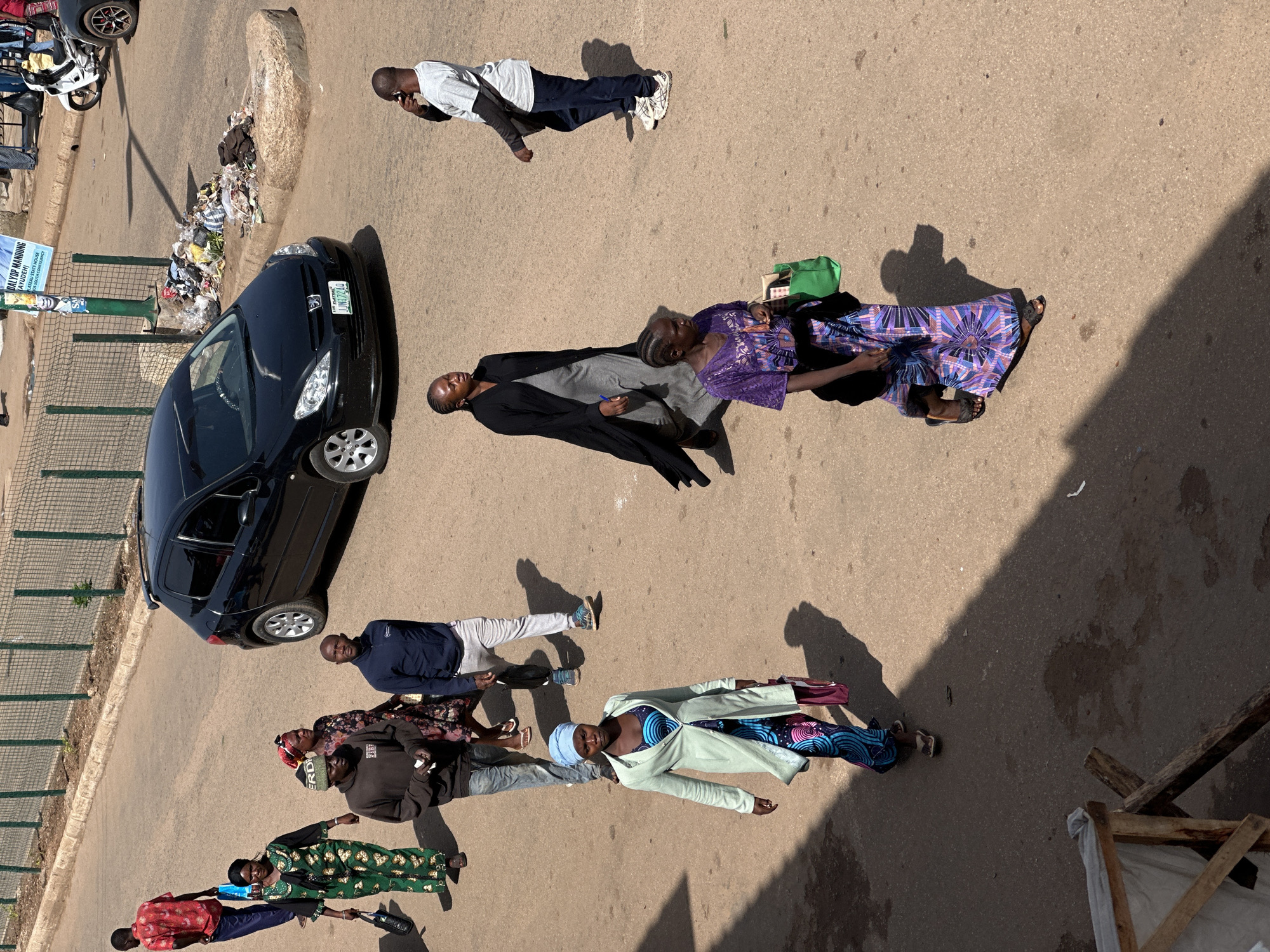 An overhead medium shot captures a street scene in Bukuru, Nigeria, with several individuals walking on an asphalt surface. A black Peugeot 308 hatchback is parked on the right, with a white motorcycle visible behind it. A pile of trash is situated near the curb on the left, adjacent to a green metal fence.
In the foreground, a woman wearing a light green coat over a blue and black patterned dress, with a blue headscarf, walks towards the camera. She carries a burgundy bag in her right hand. To her right, another woman in a purple and blue patterned dress walks away from the camera, holding a green bag and wearing dark sandals.
Further back, a man in a dark blue jacket and light grey pants walks away from the camera. To his left, a man in a brown hooded sweatshirt and blue jeans walks towards the camera, holding a white item. A woman in a green and black patterned outfit walks to the left, carrying a black bag. Another woman in a floral print dress is partially visible behind her. A man in a red short-sleeved shirt and blue jeans walks away from the camera in the upper left.
In the mid-ground, a man in a black robe over a grey dress walks away from the camera. To his right, a man in a white t-shirt and blue jeans walks towards the camera, holding a phone to his ear. The environment appears to be a sunny day, with distinct shadows cast by the people and the car. The road surface is a light brown asphalt. A white advertisement poster is attached to the green fence in the background.