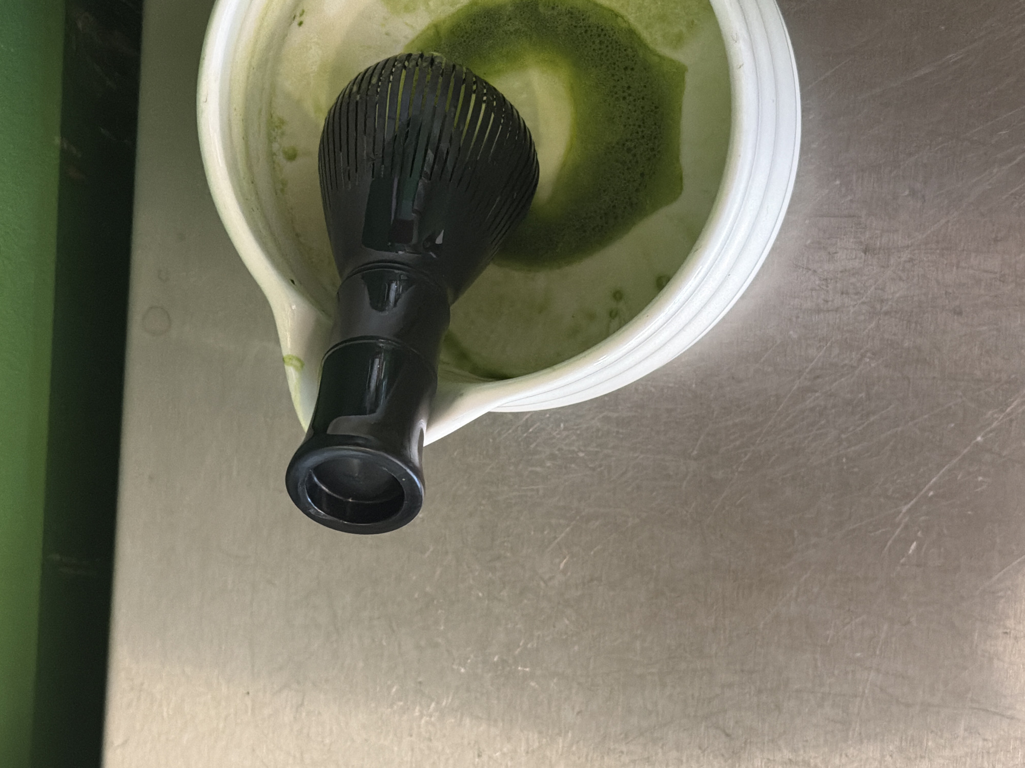 A slightly aerial, high-angle shot shows a bowl of matcha green tea with a black whisk placed inside it, set on a metallic surface. The bowl is white with a thick rim and a small spout on one side. Inside the bowl, the matcha tea has a frothy appearance, with some of the powder still undissolved near the center. The whisk is made of dark material and has a bulbous handle. The metallic surface surrounding the bowl appears to be stainless steel, marked with scratches and some water spots. The photo is taken from an oblique angle, with a green panel partially visible on the left side of the image. There is no indication of the city of Sofia, Bulgaria, or any people present in the scene.