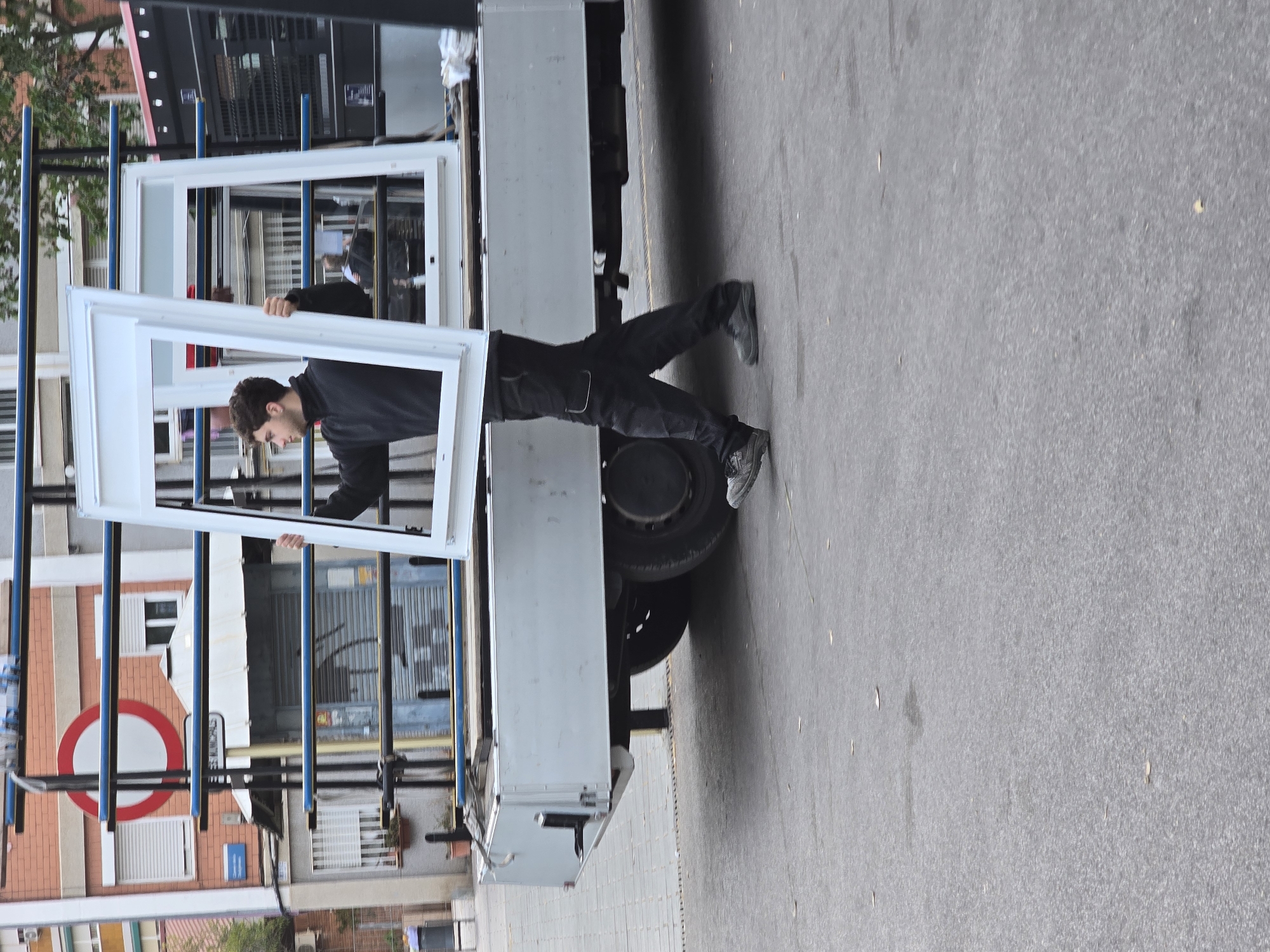 The image captures a scene of a worker handling large window frames on a street in Cornellà de Llobregat, Spain. A young man, dressed in a dark long-sleeved shirt, dark pants, and dark athletic shoes, is positioned with his torso inside the cargo area of a light grey truck. He is actively engaged in moving or securing a large white, possibly PVC or aluminum, window frame. Another similar frame is visible standing upright in the truck's bed, suggesting a delivery or installation task.
The truck, which appears to be a flatbed or open-top model, is parked on the asphalt road surface. Blue vertical poles are visible on the truck, likely used to secure its load. The background features several urban buildings. One prominent building has a brick facade with horizontal banding, and multiple windows with roller shutters. A red and white circular road sign, indicative of a "No Entry" restriction, is clearly visible in the mid-ground. Other buildings with various architectural styles and details, including windows and possibly garage doors, line the street. The scene is brightly lit, suggesting it is daytime with clear or lightly overcast weather. No specific time of day or emotional states are discernible. There is no clearly legible text on the buildings or the truck. The overall impression is that of construction-related work or material transport taking place in an urban environment.