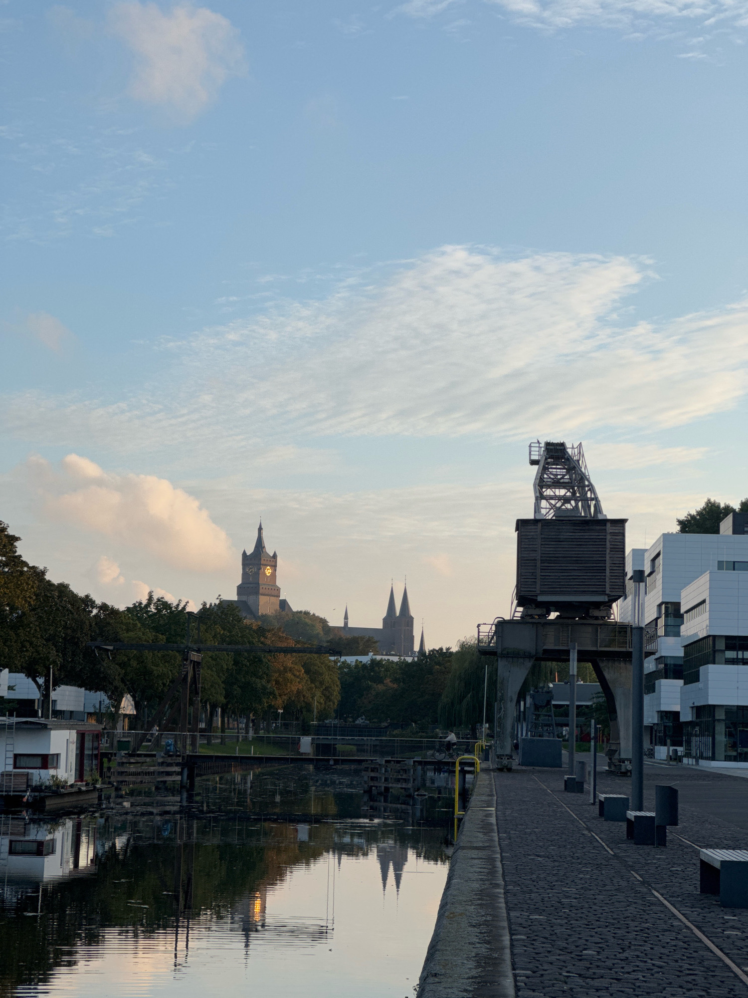 The image depicts a harbor scene in the city of Cleves, Germany, likely in the early morning or late afternoon given the soft light. In the foreground, a calm body of water reflects the sky, trees, and buildings. A white houseboat with red accents is moored on the left side of the water. A cobbled walkway runs along the right bank of the harbor, with several dark-colored benches positioned along it.
Dominating the mid-ground is a large, old-fashioned industrial crane, constructed from dark metal beams. Beyond the crane, a modern white building with dark accents stands to the right. In the distance, a prominent church with a tall, dark spire and a clock face is visible, surrounded by lush green trees. Another church with two spires can be seen further to the left in the distance. The sky is a pale blue, with scattered white, wispy clouds. The overall impression is one of urban tranquility and industrial heritage coexisting. There are no people clearly visible in the image.