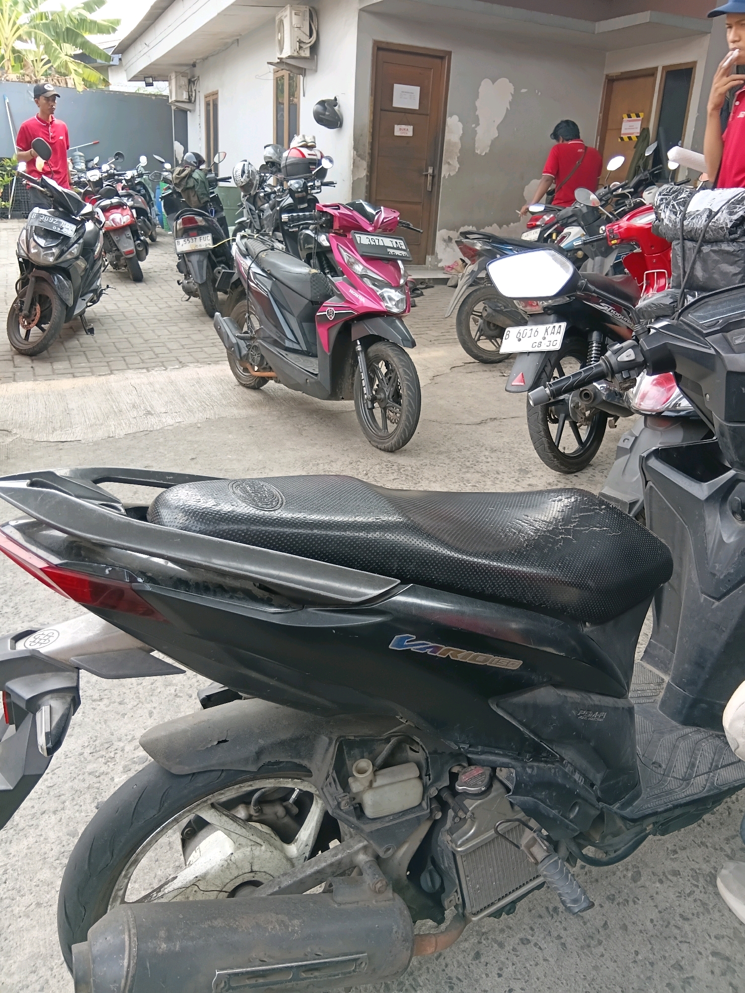 The image depicts a collection of motorcycles parked outdoors, likely in a city environment in Bekasi, Indonesia. In the foreground, a black motorcycle's seat is prominently featured, showing signs of wear and tear. Several other motorcycles, in various colors including pink and red, are visible behind it, parked in a seemingly disorganized fashion. Some individuals are present, dressed in red shirts, suggesting they might be employees or delivery personnel. One person, wearing a red shirt and a black cap, is standing and holding a round object, possibly a helmet. Another individual in a red shirt is seated on a motorcycle in the background. The setting appears to be a paved area in front of a building with light-colored walls and several doors. The overall scene suggests a busy area where motorcycles are a common mode of transport.