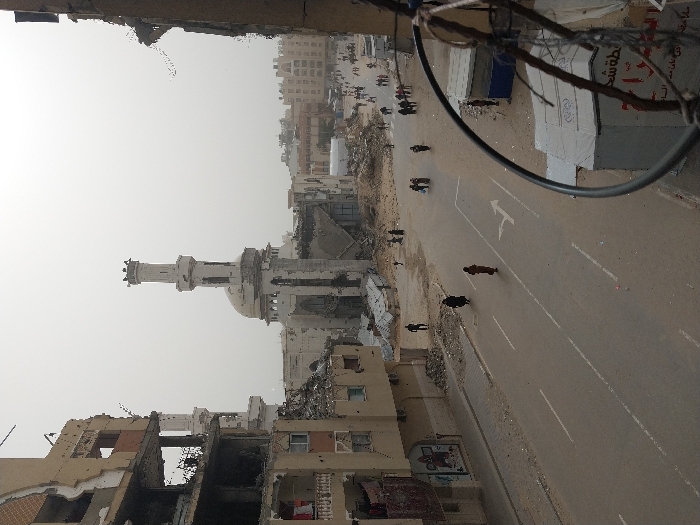 The image depicts a heavily damaged urban landscape in Khan Yunis, Palestinian Territory, under an overcast, hazy sky. The central feature is a large mosque with a prominent minaret and dome, both showing significant structural damage, including visible erosion and missing sections of masonry, particularly around the dome's base. The minaret features a distinct octagonal upper section with a crenellated top.
To the immediate left of the mosque, a multi-story building is extensively damaged, with multiple floors partially collapsed, exposed interiors, and rebar protruding from shattered concrete. Piles of rubble, consisting of concrete, rebar, and other debris, are scattered across the ground in front of the mosque and along the sides of the main paved road that runs diagonally from the bottom left towards the background. The road itself has visible white lane markings, including a solid double line and a painted white right-turn arrow.
Numerous individuals are present throughout the scene. In the mid-ground, a few people, predominantly wearing dark clothing, are walking on the road and sidewalks. A woman in a reddish-brown garment walks near the center of the road. Further in the distance, larger groups of people are congregated or walking, suggesting ongoing activity despite the destruction.
On the far right, partially obscured by a dark, circular metal object and exposed wires in the immediate foreground (indicating the vantage point is from a damaged structure), another building edge is visible, also showing signs of destruction. A white, tent-like structure with Arabic script is also present on the right side of the road in the mid-ground. The background reveals additional urban structures, some appearing less damaged than the foreground buildings, but the overall environment signifies extensive conflict damage and debris.