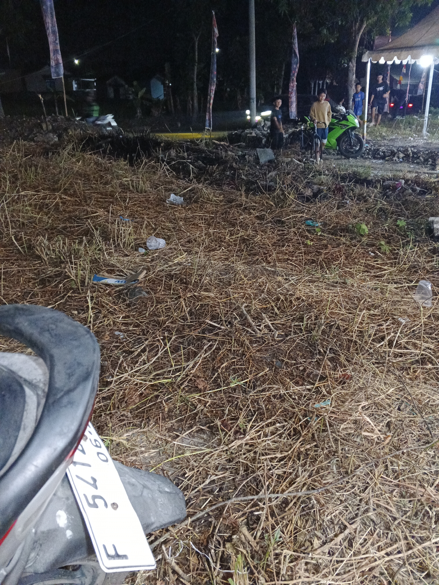 This is a medium shot, low-angle, outdoor, night photograph taken in Sukabumi, Indonesia. The foreground is dominated by dry grass and debris, with the rear of a motorcycle, including its license plate reading "F 5417 VG 06", visible in the bottom left. In the middle ground, several young men are gathered around a bright green sport motorcycle. The background shows trees, some structures that appear to be houses, and a white tent with lights, suggesting an outdoor gathering or event. The scene is dimly lit, characteristic of nighttime conditions.