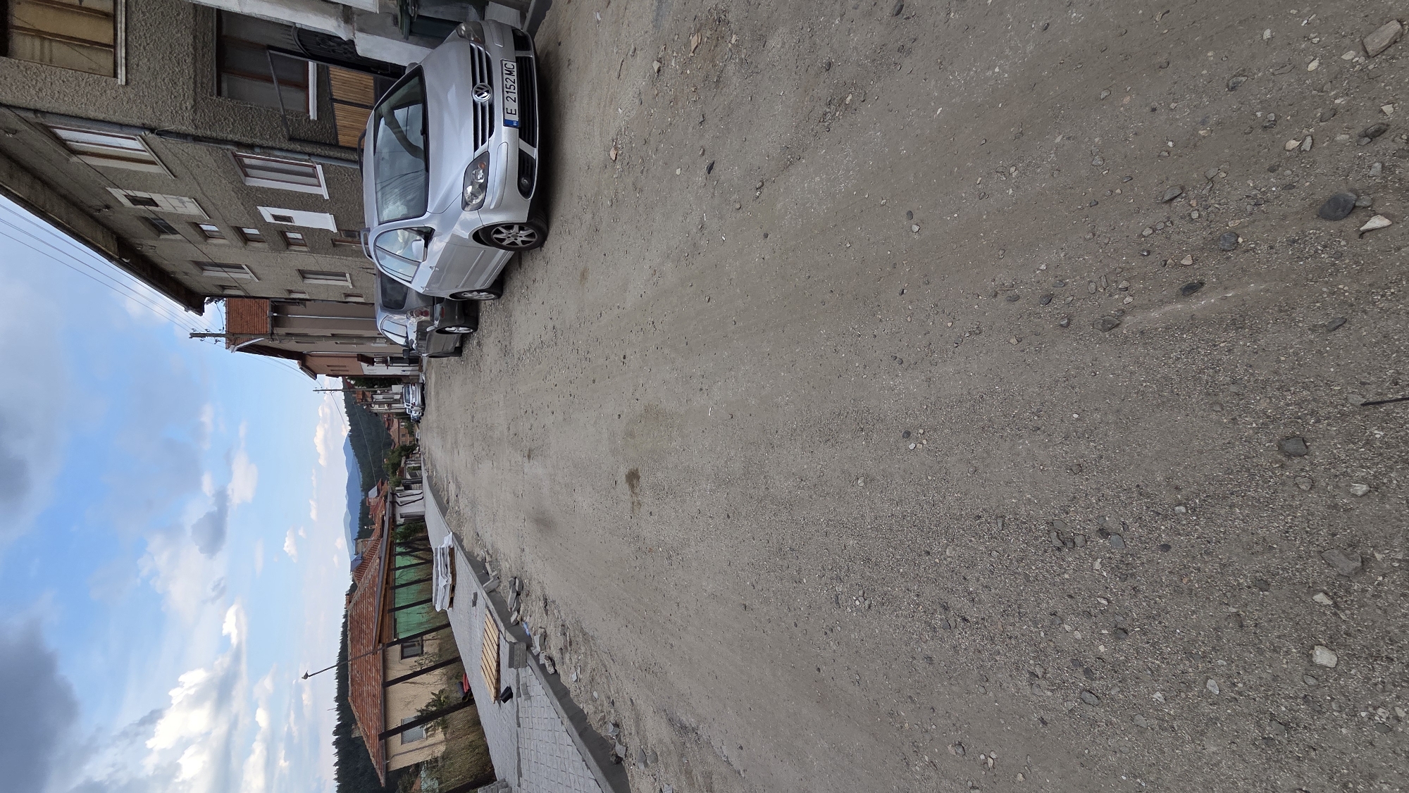 The image shows a rural street under construction in the city of Bania, Bulgaria. The street is unpaved and appears to be covered in gravel and dirt. On the left side of the street, there is a newly laid sidewalk with paving stones. A stack of wooden pallets is also visible on the sidewalk. Several cars are parked along the right side of the street, including a silver Volkswagen Golf. Buildings line both sides of the street, and the sky above is partly cloudy. The overall impression is one of ongoing infrastructure development in a small town.