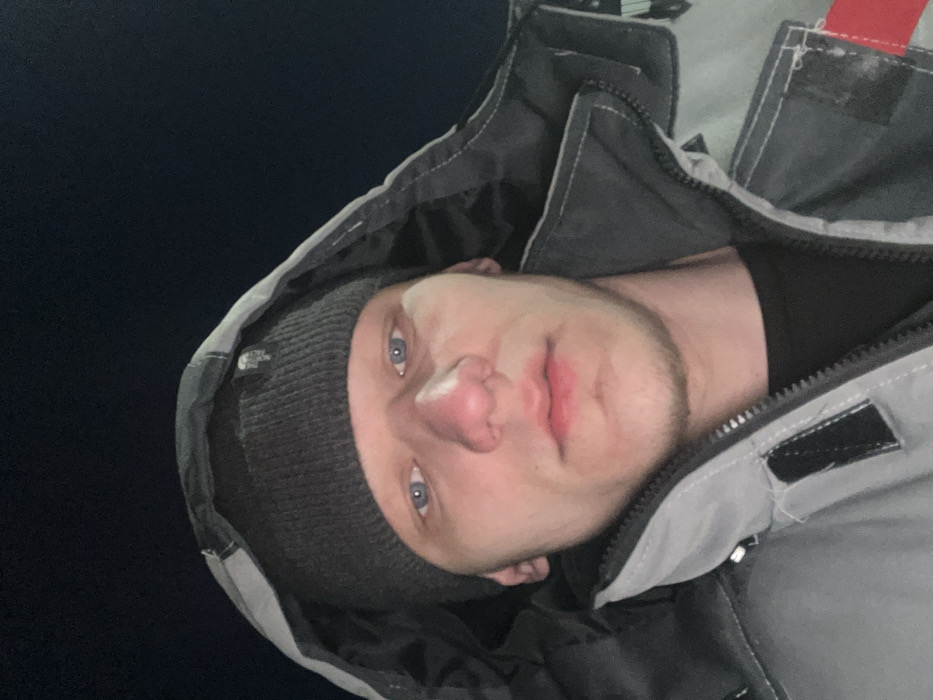 A close-up, upward-angled selfie depicts a young male individual located in the city of Залесная, Russia. The subject has light blue eyes, a pronounced nose, and light stubble visible on the chin and jawline. He is wearing a dark grey knit beanie with a small logo on the front and a grey and dark grey hooded jacket. The jacket features a zipper closure, lighter grey sections on the upper torso and arms, and a red/orange strip visible on the lower right sleeve. A black t-shirt is visible beneath the jacket. The individual maintains a neutral expression, looking directly at the camera. The background is uniformly dark, indicating a nighttime setting with no discernible environmental features.