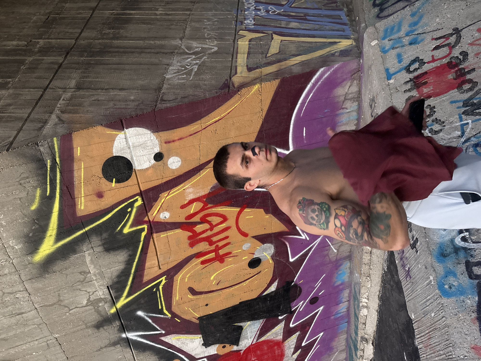 A young, shirtless man with a short haircut and a black band-aid on his nose stands in front of a graffiti-covered concrete wall. He has tattoos on his left arm, including a skull wearing a crown, and he is holding a dark red cloth in his right hand. The graffiti on the wall is colorful and abstract, with bold lines and shapes. The man is wearing white pants. The scene appears to be in an outdoor urban setting, possibly under a bridge or overpass, given the concrete structure and the nature of the graffiti. The location is Varna, Bulgaria, specifically the area beneath the Asparuhovo Bridge.