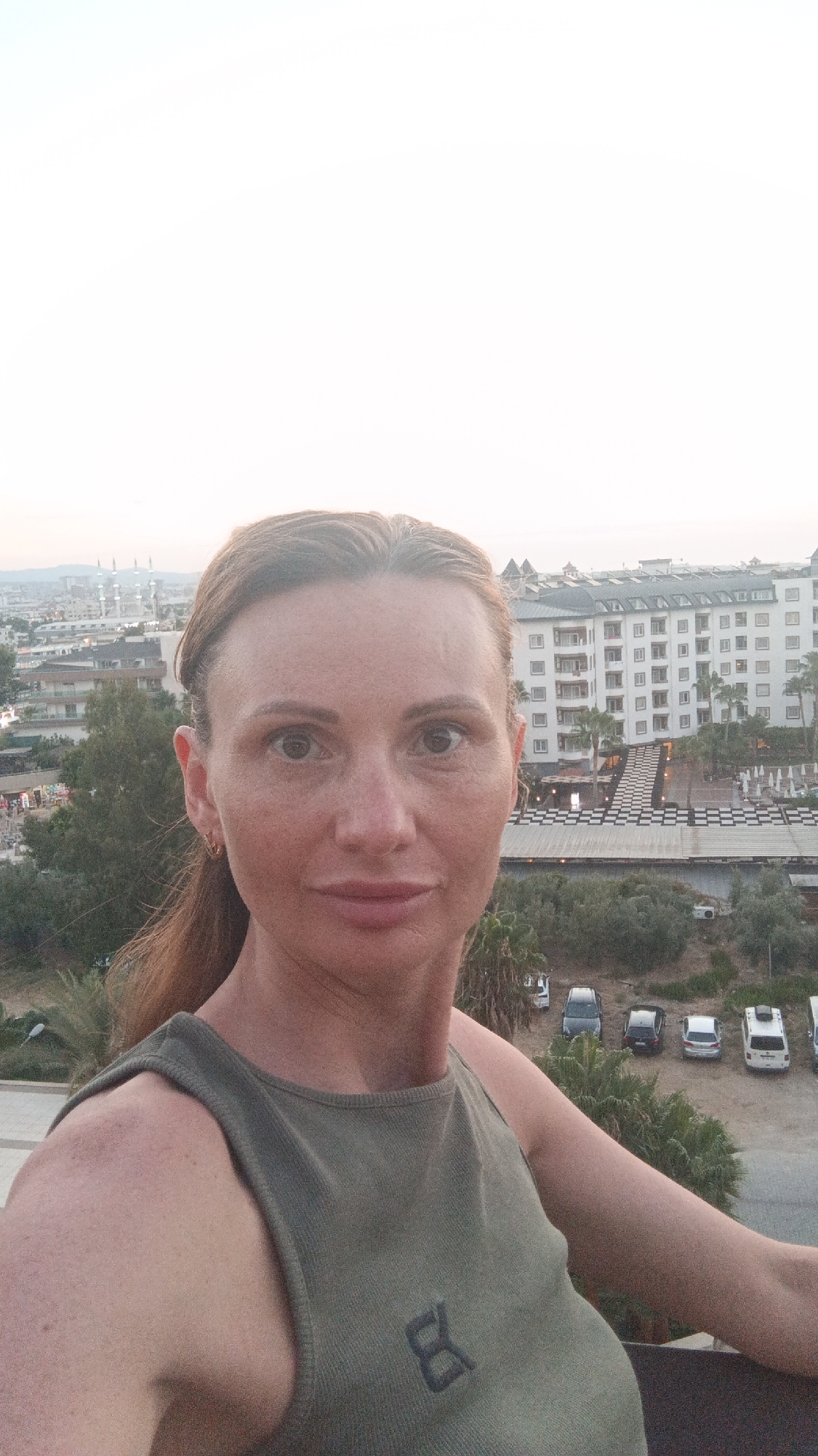 A woman with light skin and brown hair pulled back is taking a selfie from a high vantage point. She is wearing a sleeveless, olive-green tank top with a black logo on the left chest. Her expression is neutral. In the background, a city skyline is visible, featuring low-rise buildings and several minarets, characteristic of Alanya, Turkey. A large, white resort-style building with multiple floors and balconies is prominent on the right. Below the resort, there are palm trees, an outdoor seating area with checkered flooring, and a parking lot with several cars. The sky is overcast and pale. The overall scene suggests an urban environment in Alanya, Turkey, during daylight hours, possibly late afternoon or early evening due to the soft light.