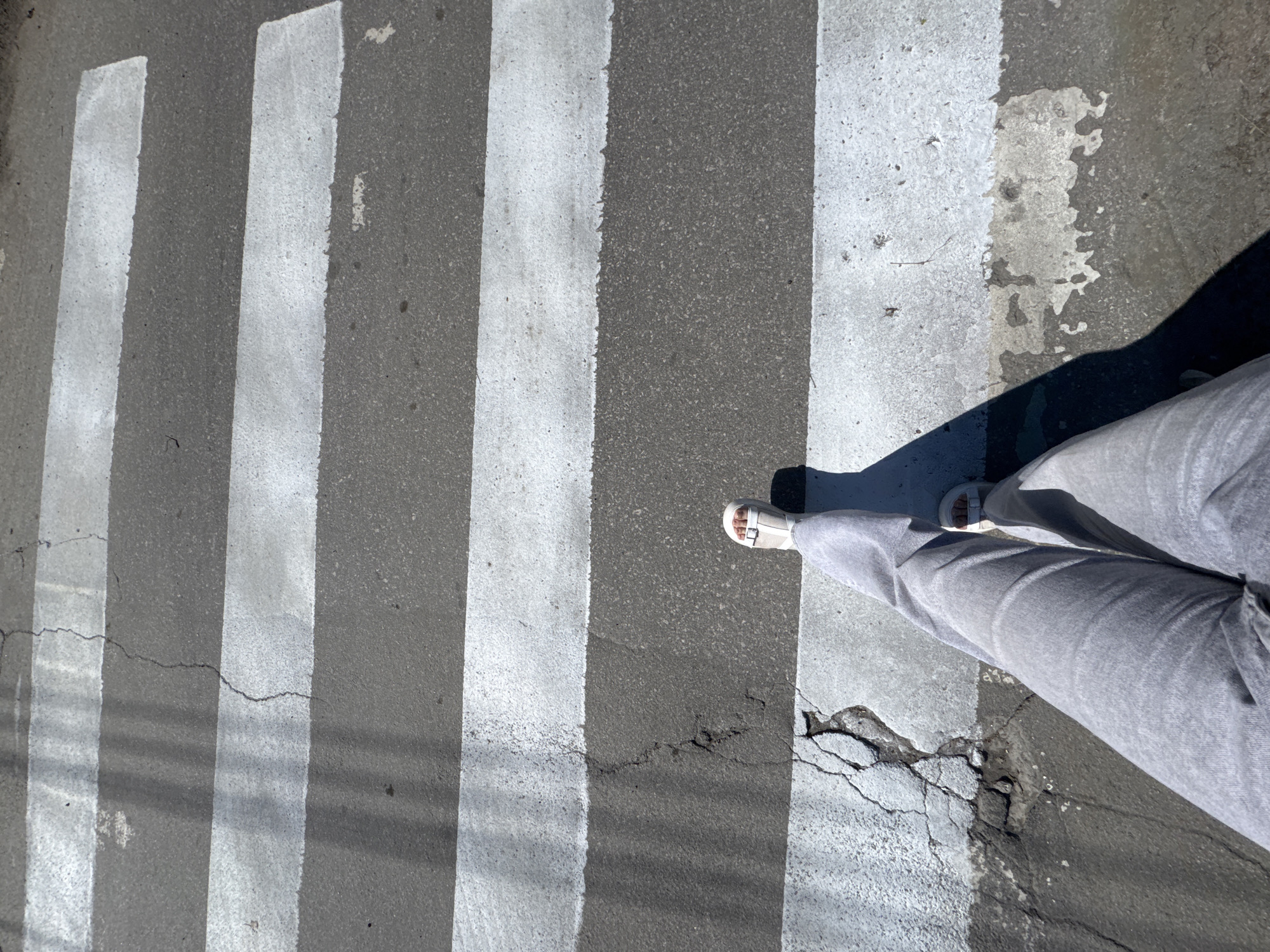 A person is standing on a crosswalk in an urban environment. The crosswalk consists of alternating white stripes and grey asphalt, oriented perpendicular to the direction of pedestrian traffic. The asphalt shows signs of wear and cracking. The person is wearing grey pants and white sandals, with their shadow cast towards the right side of the frame. The location is Unknown, Unknown.