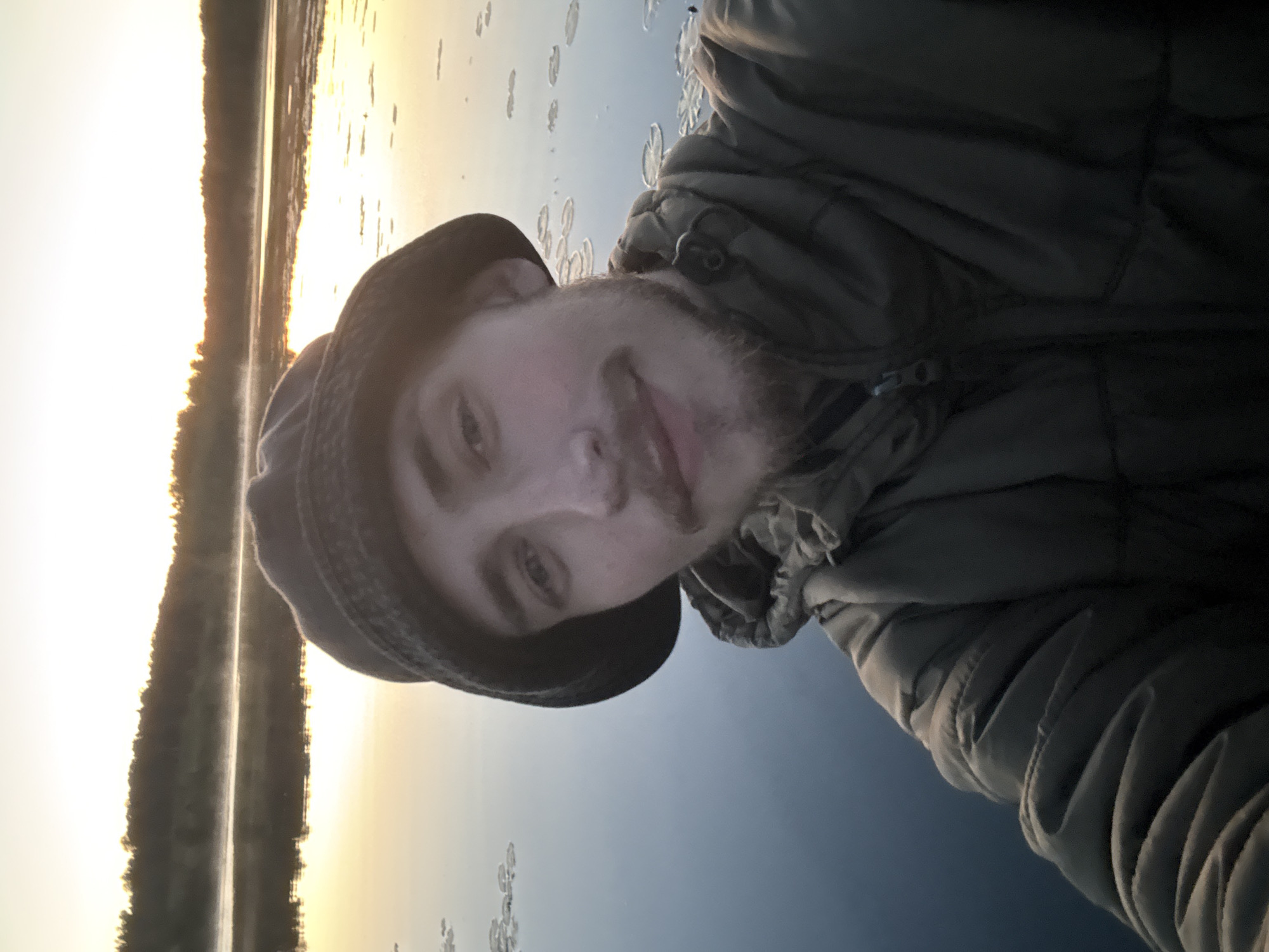 A man is taking a selfie by a lake at sunset. He is wearing a dark bucket hat and a green puffer jacket. The lake reflects the warm colors of the sunset, and the distant shoreline is lined with trees. Some lily pads are visible on the surface of the water. The image appears to be taken from a boat on the water. The location is the city of Rogovoe, Russia.
