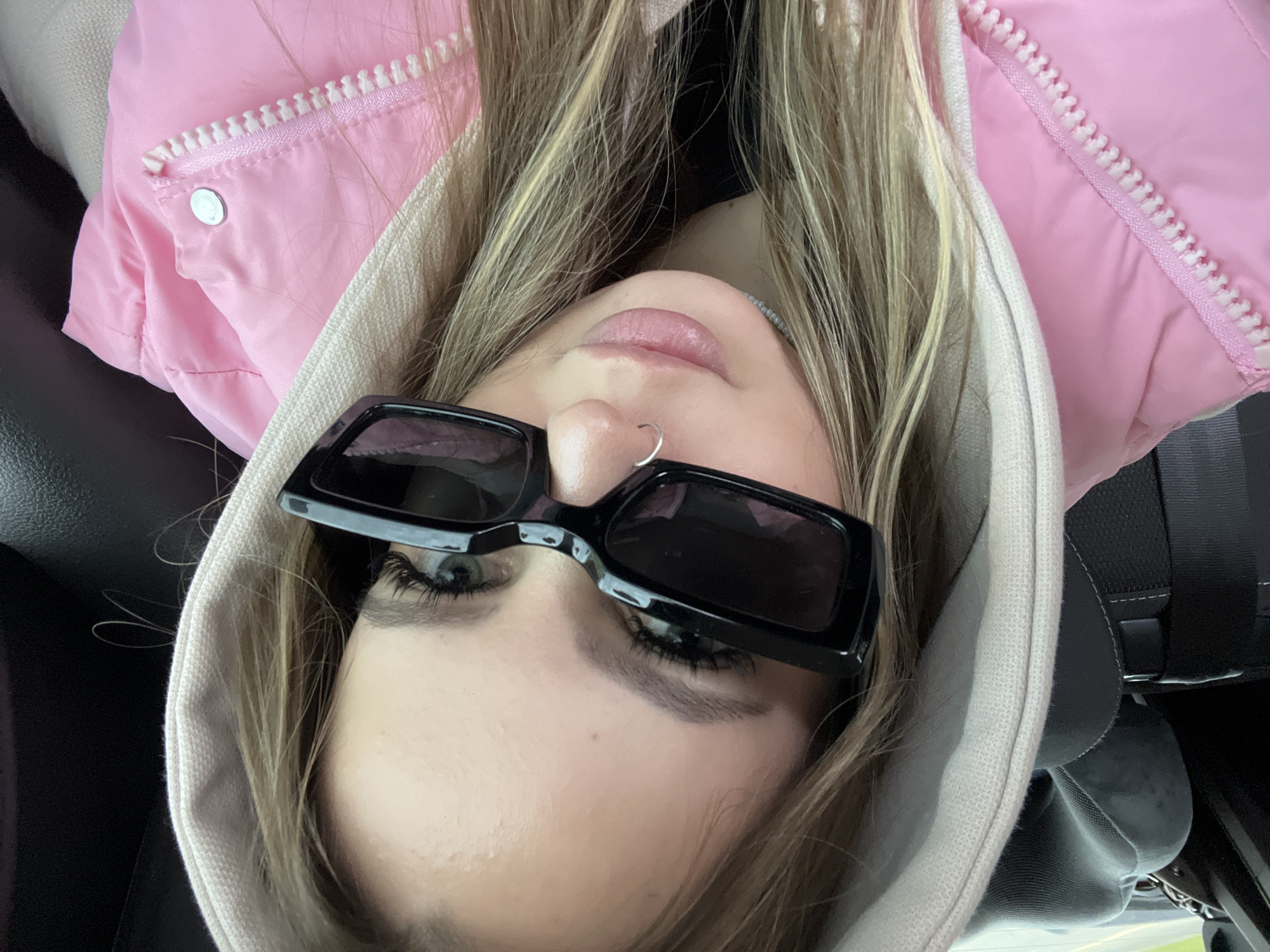 An overhead, close-up shot captures a young adult, likely female, positioned upside down from the viewer's perspective, possibly taking a selfie. Her long, straight blonde and light brown hair cascades around her face. She is dressed in a vibrant pink puffer jacket, featuring a white zipper and a silver snap button on the shoulder, paired with a light beige or off-white hoodie.
Her face is largely obscured by stylish black, rectangular sunglasses, though one light-colored eye with prominent dark eyelashes is visible above the left lens. Her eyebrows are well-defined. A small, silver hoop ring pierces her right nostril. Her lips are slightly parted, showcasing a natural pink shade. The expression appears neutral.
The setting is an interior space, specifically a car, evidenced by the dark upholstery visible to the left and elements of a car seat or seatbelt system on the right. No external environment is visible. The image conveys no specific time of day or weather. No text is visible within the frame. This scene takes place in Chituk, Russia.