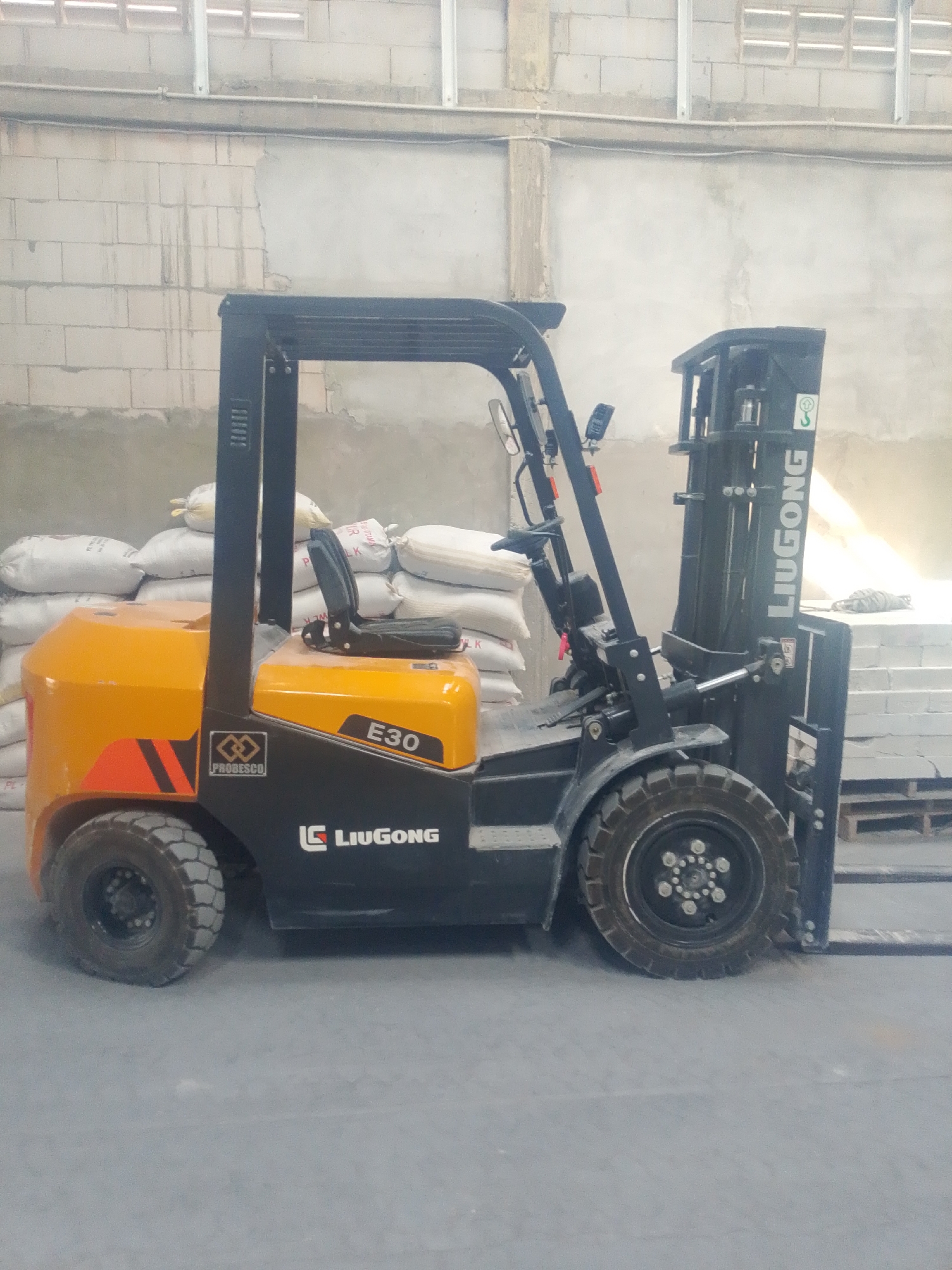 A forklift, identified by "LIUGONG" and "E30" branding, is parked indoors. The forklift is yellow and black and has a protective cage over the driver's seat. Piles of white sacks are stacked behind the forklift. The background consists of a concrete block wall and a concrete floor. The scene appears to be a warehouse or storage facility. The image has a bright, natural light source coming from the right side. There are no people visible in the image. The approximate location of this facility in Banjarmasin, Indonesia, is unknown, as the image does not contain any specific identifying features of that city.