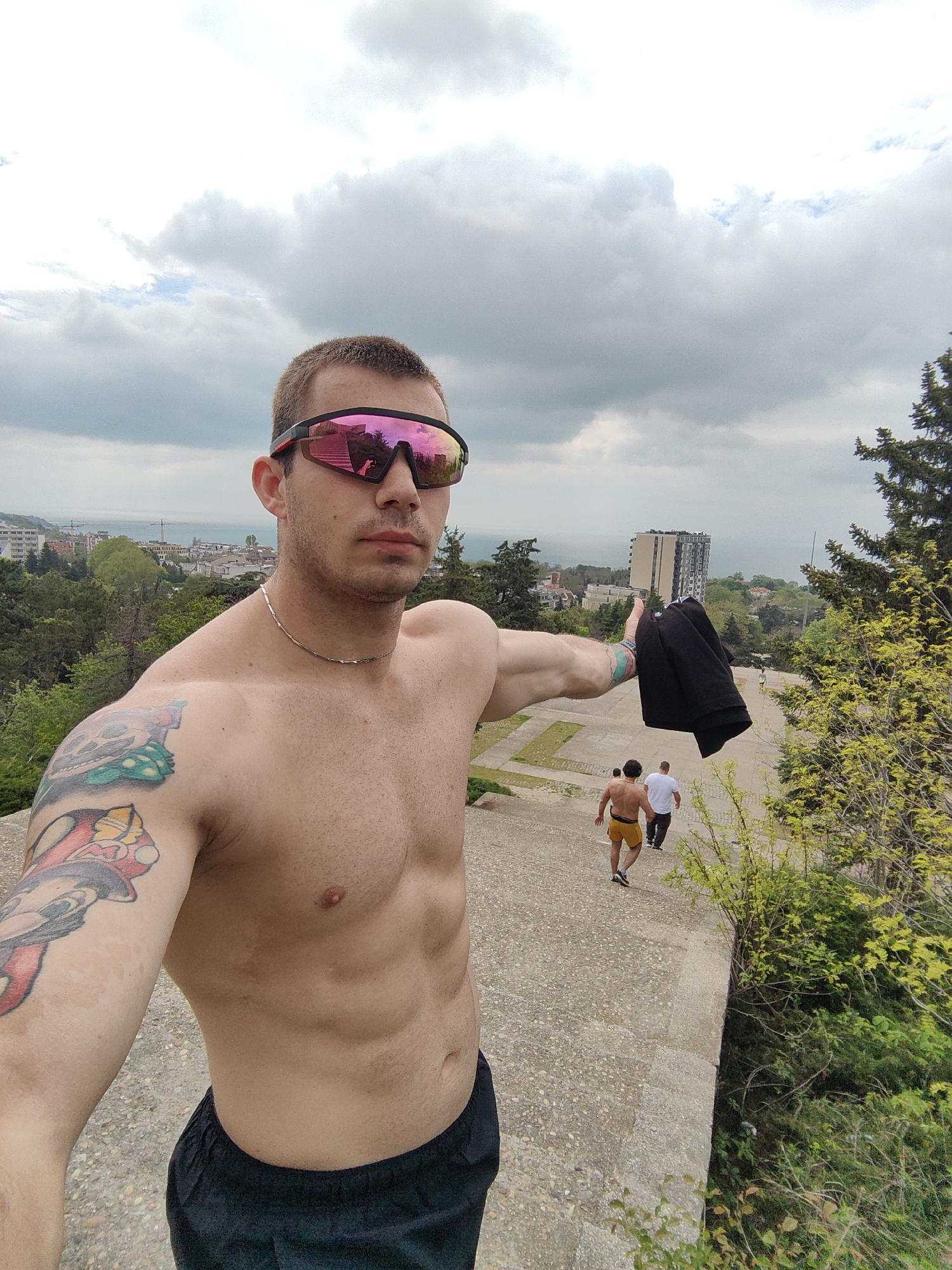 A shirtless man with pink sunglasses takes a selfie outside. He has a tattoo on his right arm and is wearing a silver necklace. He is holding a black jacket or shirt in his right hand, pointing towards a building in the background. Two men walk down the steps behind him. The background includes trees, buildings, and a cloudy sky. The location is Varna, Bulgaria, as indicated by the architecture and landscape.