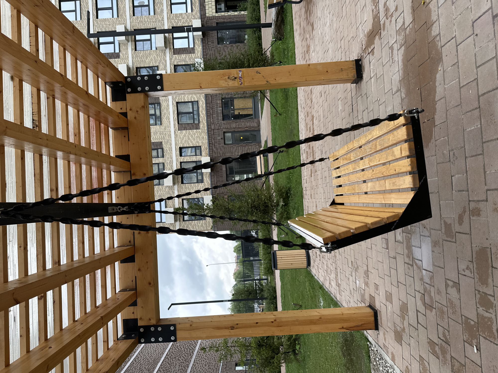 A wooden swing set with a slatted wooden seat is suspended from a wooden pergola with a translucent corrugated roof. The swing set is situated on a paved area of reddish-brown bricks. To the left of the swing set is a grassy area with small evergreen trees and a wooden trash receptacle. In the background, there is a multi-story brick building with numerous windows and balconies, characteristic of urban residential architecture in Znamya Oktyabrya, Russia. The sky is overcast, suggesting a cloudy day, and there are puddles on the ground, indicating recent rain. There are no people or animals visible in the scene.