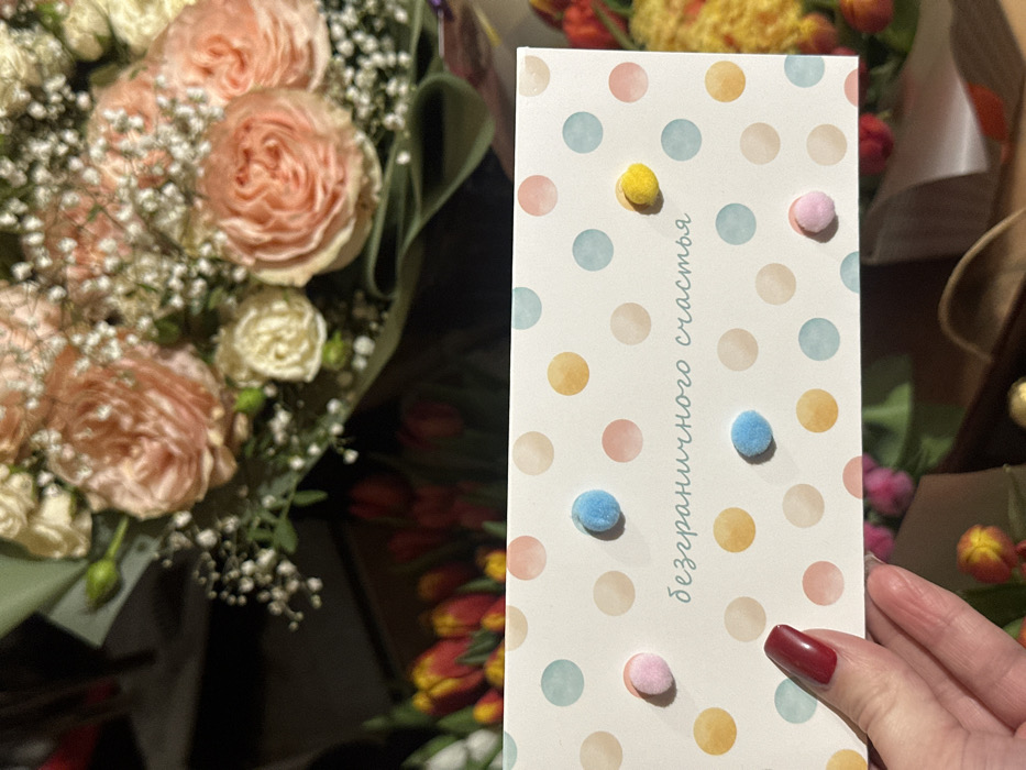 A close-up image taken in Базарово, Russia, depicts a left hand with dark red manicured fingernails holding a white greeting card in the lower-left quadrant. The card features a pattern of watercolor-style polka dots in various pastel shades (pink, blue, yellow, beige) and is adorned with small, fuzzy pom-poms in light pink, light blue, and yellow. The Russian text "безграничного счастья" (unlimited happiness) is printed in teal script on the card. In the blurred background, a large bouquet of light peach or pink roses mixed with white baby's breath, wrapped in a dark green paper or fabric, occupies the upper portion of the frame. Additional out-of-focus floral arrangements, including red and yellow tulips, are visible in the background, suggesting a flower shop or similar retail environment.
