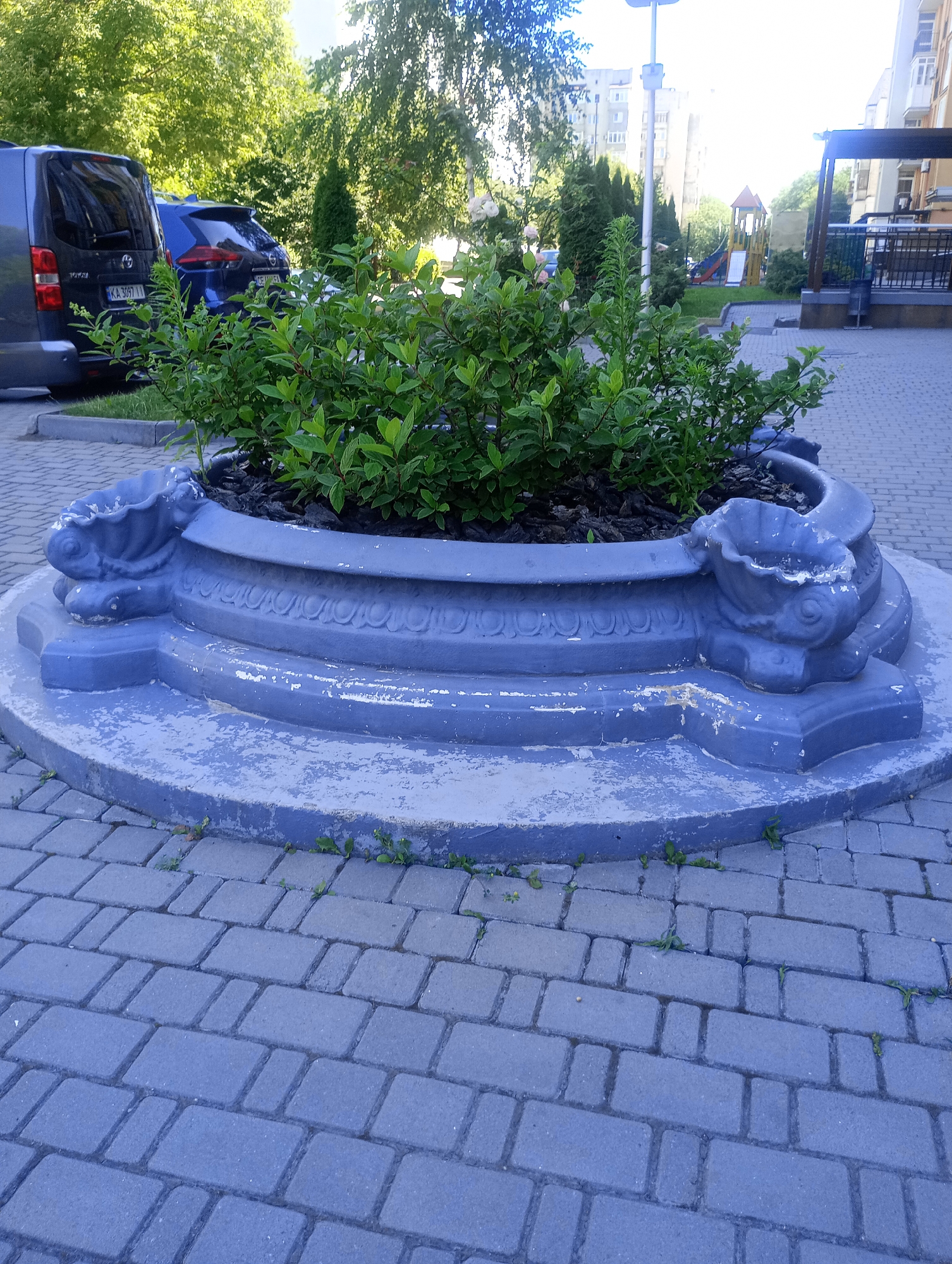 A concrete planter with ornate, fish-like sculptures on either side holds a dense cluster of green leafy plants. The planter is painted a faded blue, with visible signs of wear and chipping paint on its surface. It sits on a circular concrete base, surrounded by a paved walkway made of gray interlocking bricks.
In the background, a dark blue van is parked, and beyond that, a blue car. Lush green trees line the area, casting dappled shadows. Further in the distance, a multi-story building and a playground with a slide and climbing structure can be seen. The sky is bright, suggesting it is daytime. The overall scene appears to be an outdoor urban or suburban setting, possibly a courtyard or residential area. There is no discernible activity taking place, and the image conveys a sense of stillness. License plate visible on the van reads "KA 3097 IA".