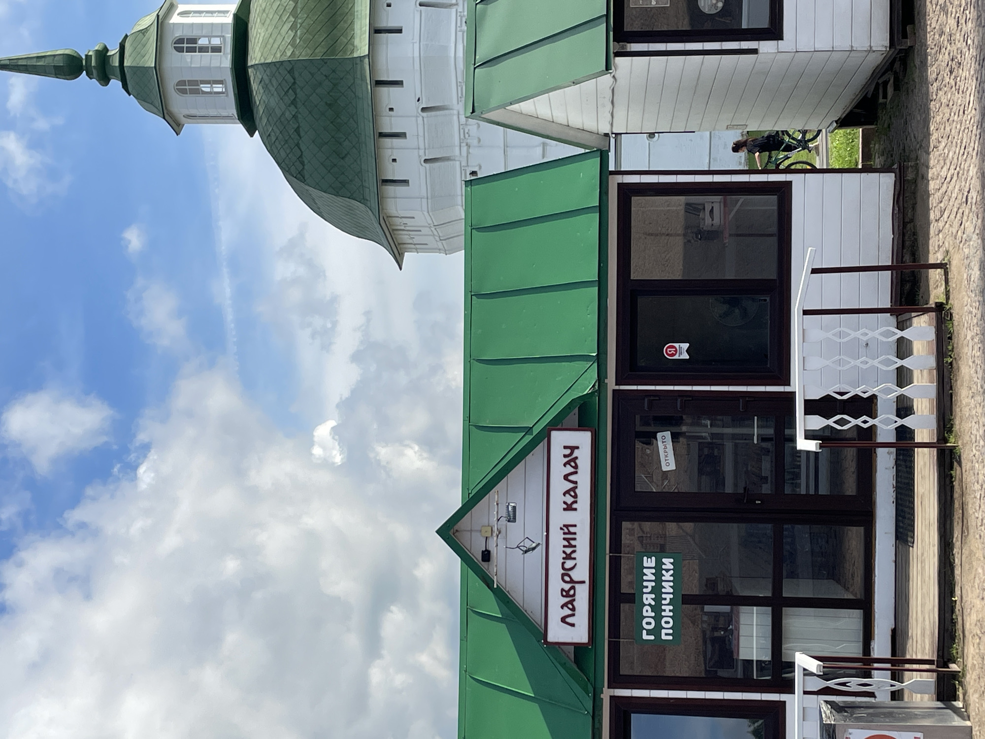 The image shows a small kiosk with a green roof and white walls, located in the city of Sergiyev Posad, Russia. A sign above the kiosk reads "ЛАВРСКИЙ КАЛАЧ" in red lettering. Below this, another sign on the glass door states "ГОРЯЧИЕ ПОНЧИКИ" in green lettering, indicating that the kiosk sells hot donuts. A smaller sign on the same door reads "ОТКРЫТО" (Open). In the background, a large white building with a green domed roof, possibly part of a monastery or church, is visible against a blue sky with scattered clouds. To the right of the kiosk, another similar structure is partially visible, and a person is seen riding a bicycle in the distance. The ground in front of the kiosk is paved with cobblestones, and there are white railings along a wooden walkway.