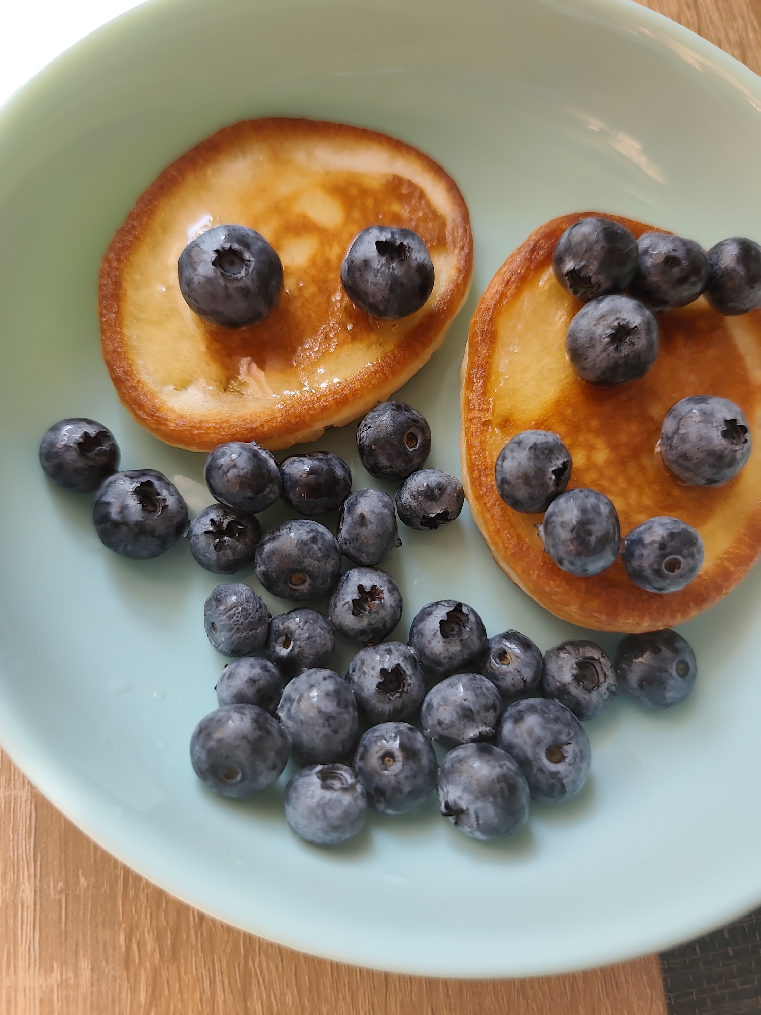A close-up, top-down shot shows two small, golden-brown pancakes arranged on a light blue, shallow bowl. Several fresh blueberries are scattered across the pancakes and the surface of the bowl. The scene is set on a wooden table, with the edge of a textured placemat visible on the right side of the image. The overall impression is of a simple, appealing breakfast. This image does not contain enough information to determine its location in Durlești, Moldova.