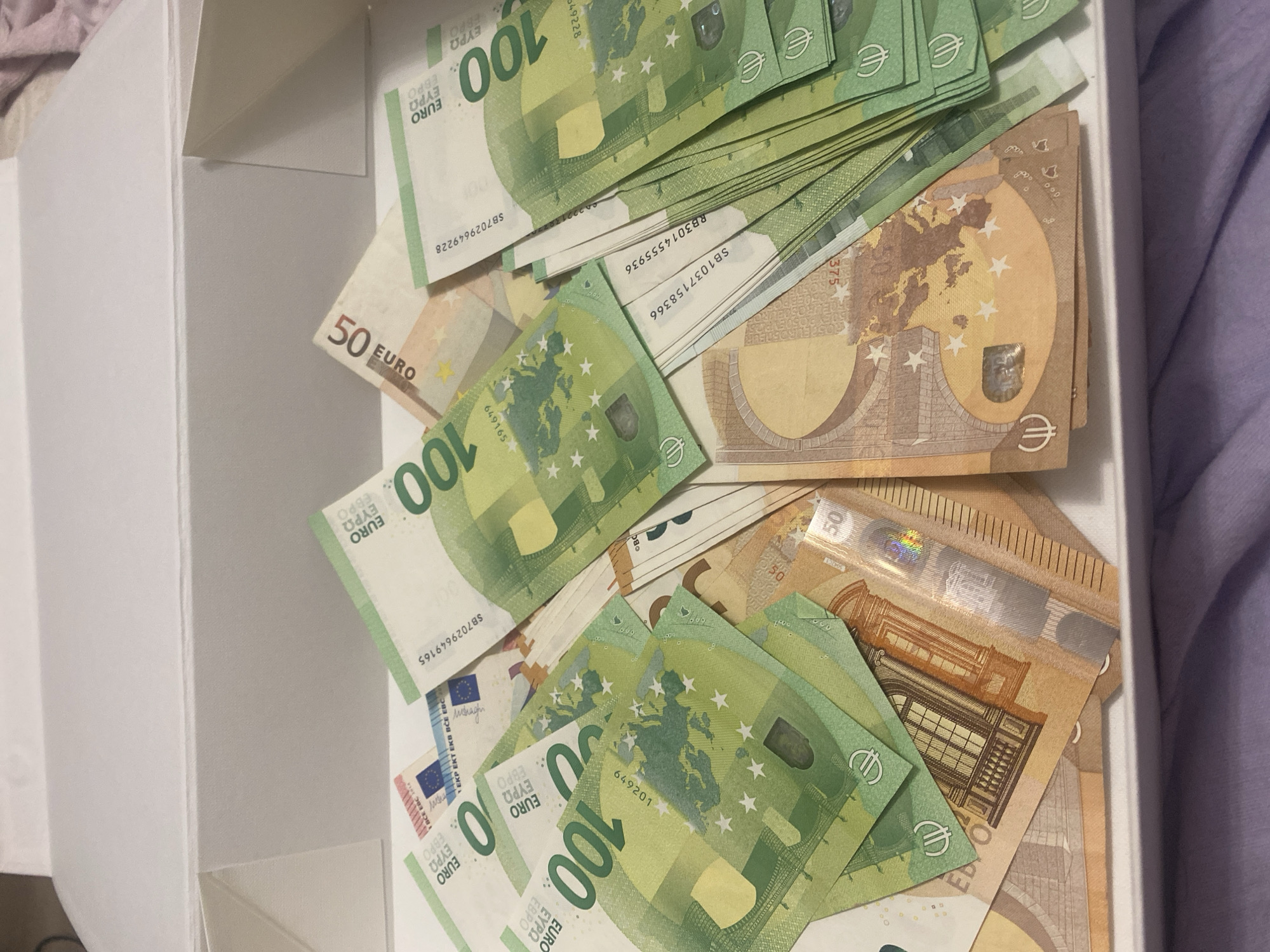 The image shows a collection of European banknotes, primarily 100 Euro notes and some 50 Euro notes, spread out inside a white box. The banknotes are arranged in a somewhat disordered manner, with some overlapping and fanned out. The lighting suggests an indoor setting, possibly a home or an office. The banknotes appear to be genuine currency. There is no indication of the location in Novi Petrivtsi, Ukraine. The image focuses on the money itself.