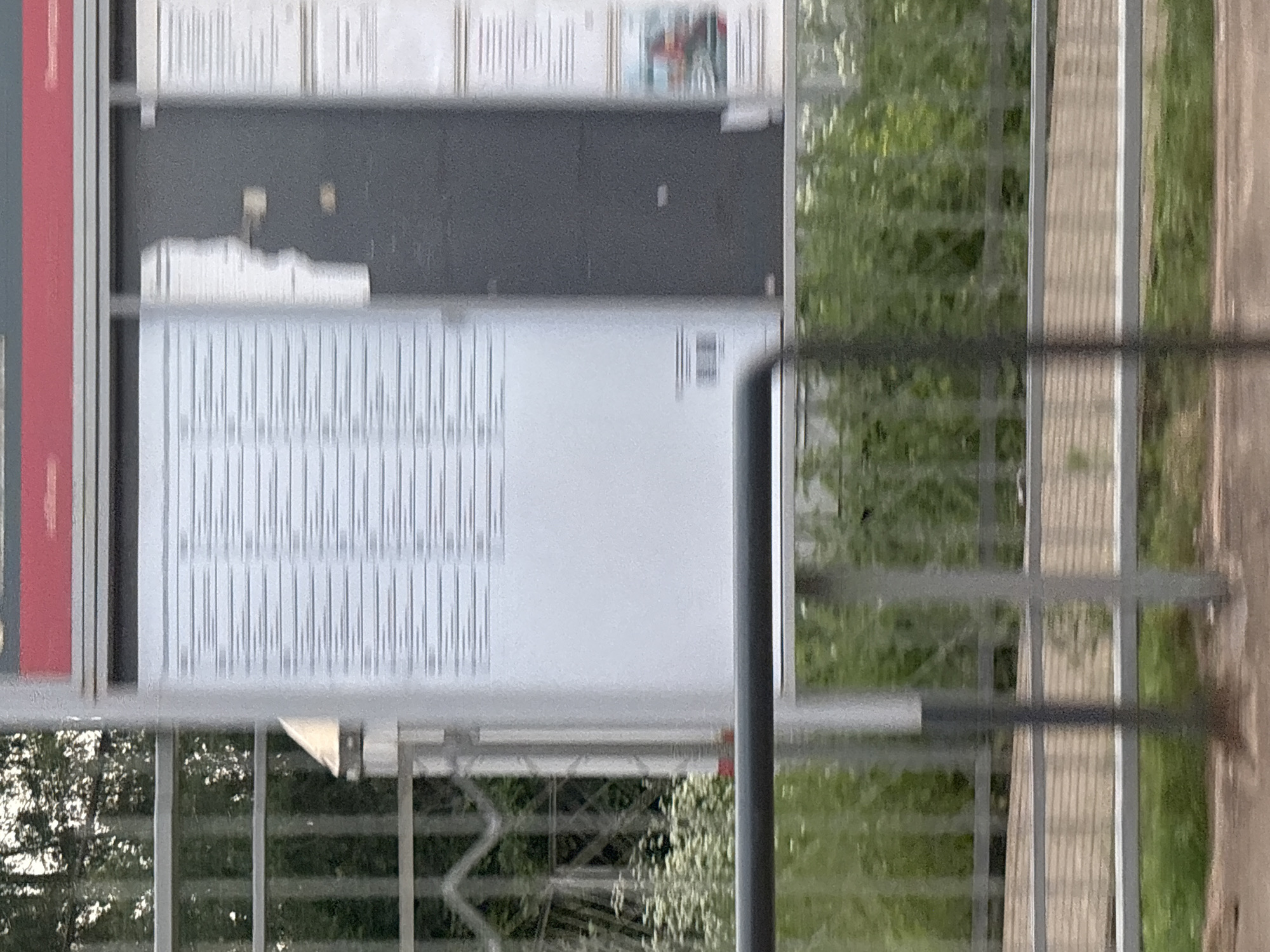 The image shows a blurred view of a timetable display at a train station in Povarovo, Russia. The timetable is mounted on a white board with some parts obscured by a dark panel. There is a red header at the top of the display. The foreground is partially obscured by a metal railing. Beyond the railing, there is lush green vegetation. The overall impression is of an outdoor setting, likely a train station platform, with the timetable serving as the central subject.