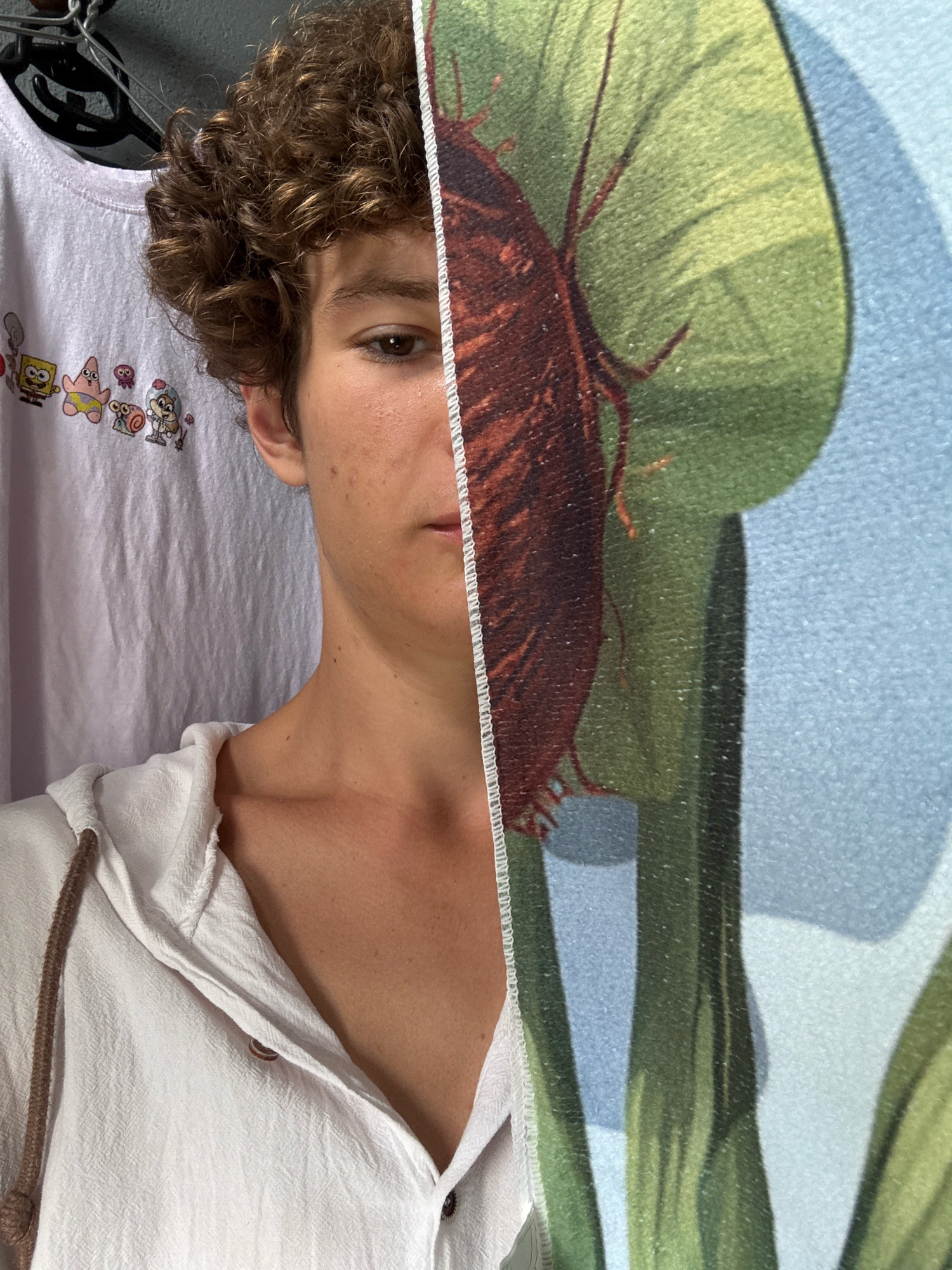 The image shows a close-up of a young person's face, partially obscured by a patterned cloth that features a large brown insect on a green leaf. The person is wearing a white hooded garment with rope drawstrings and a t-shirt with cartoon characters visible on the left side of the frame. The background appears to be a textured surface, possibly a wall or fabric. The lighting suggests an indoor setting. There is no discernible information about the location in Nha Trang, Vietnam, in this image.