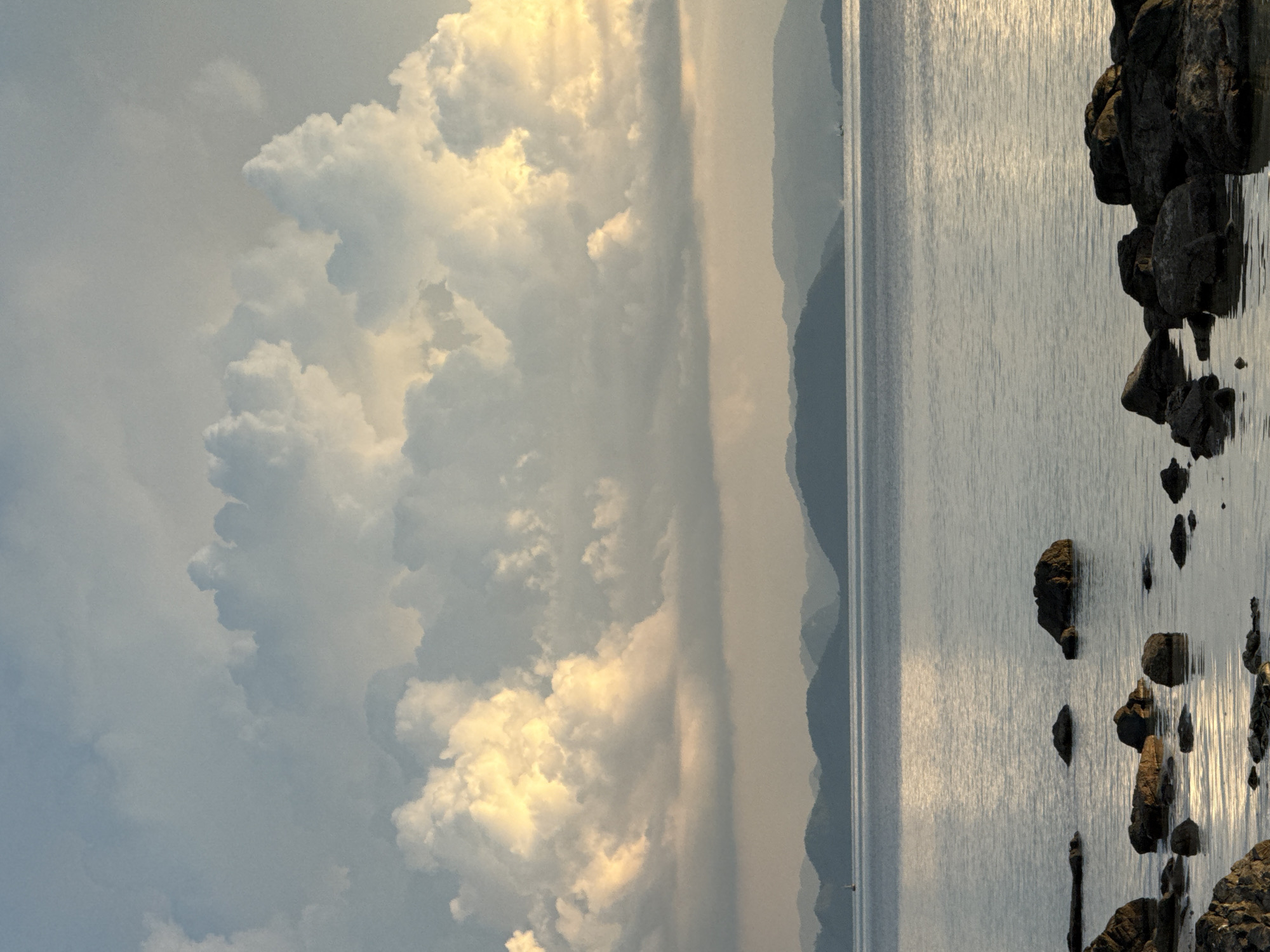 A vertical, wide shot captures a tranquil seascape with dramatic clouds at sunset. The foreground is dominated by dark, wet rocks partially submerged in calm, reflective water. The water stretches to the horizon, where a series of layered, hazy mountains recede into the distance. Above the mountains, the sky is filled with large, voluminous cumulus clouds, illuminated from behind by the setting sun, creating a warm, golden glow on their undersides, contrasting with the cooler, blue-grey tones of the upper clouds. The overall atmosphere is serene and majestic. The image is taken from the coast of Baan Pang Ka, Thailand, a location known for its picturesque bays and islands.