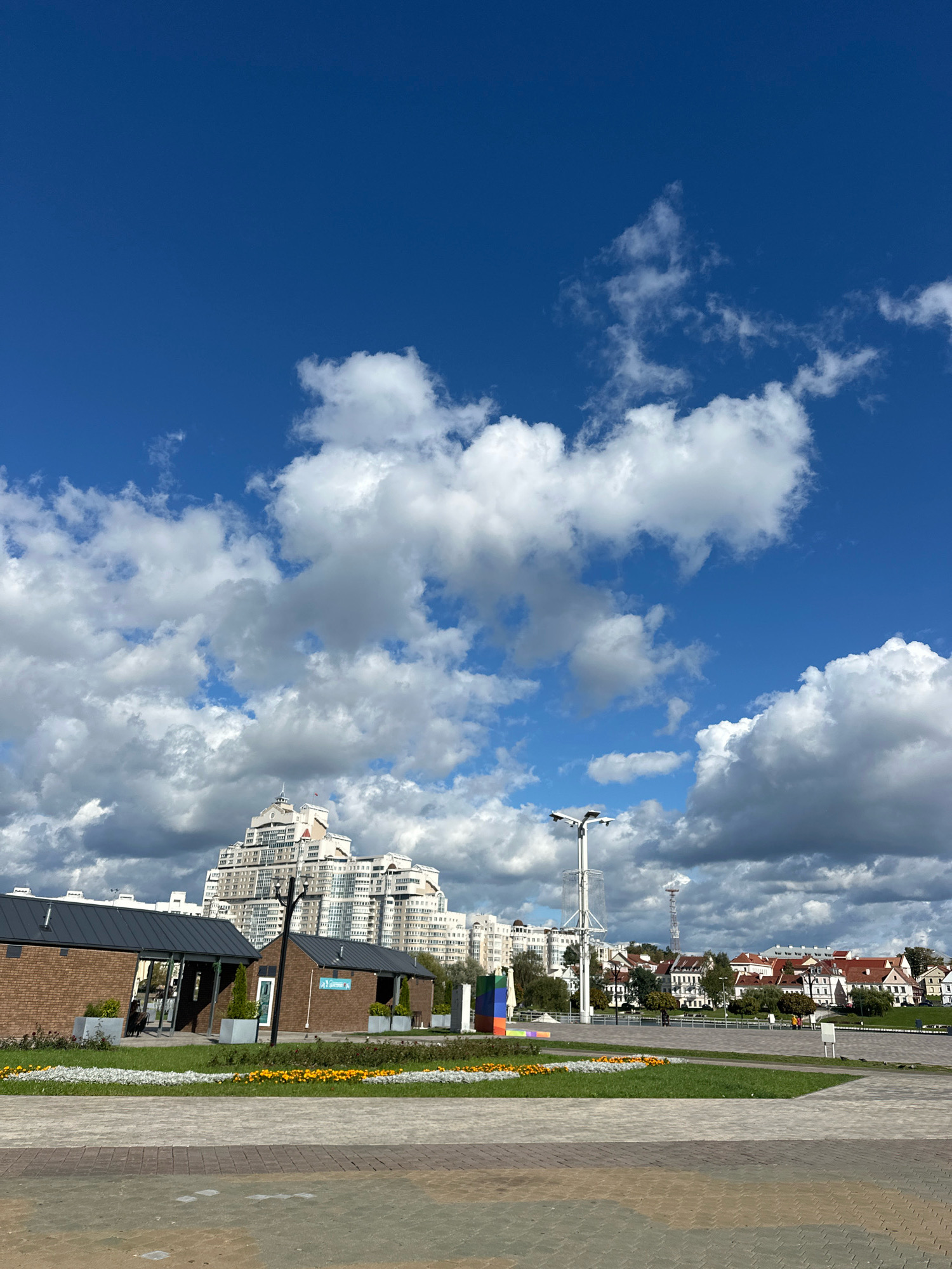 The image shows a view of the Upper Pond in Minsk, Belarus, with a bright blue sky filled with white, fluffy clouds. In the foreground, a paved walkway leads towards a green area with a flowerbed of yellow flowers. To the left, there are two small, modern brick buildings with dark roofs. Further in the background, a tall, distinctive white building with many windows dominates the skyline, characteristic of the architecture along the Upper Pond embankment. Beyond that, a row of red-roofed houses and other buildings line the horizon, and a tall, slender communication tower is visible in the distance. The scene appears calm and sunny.